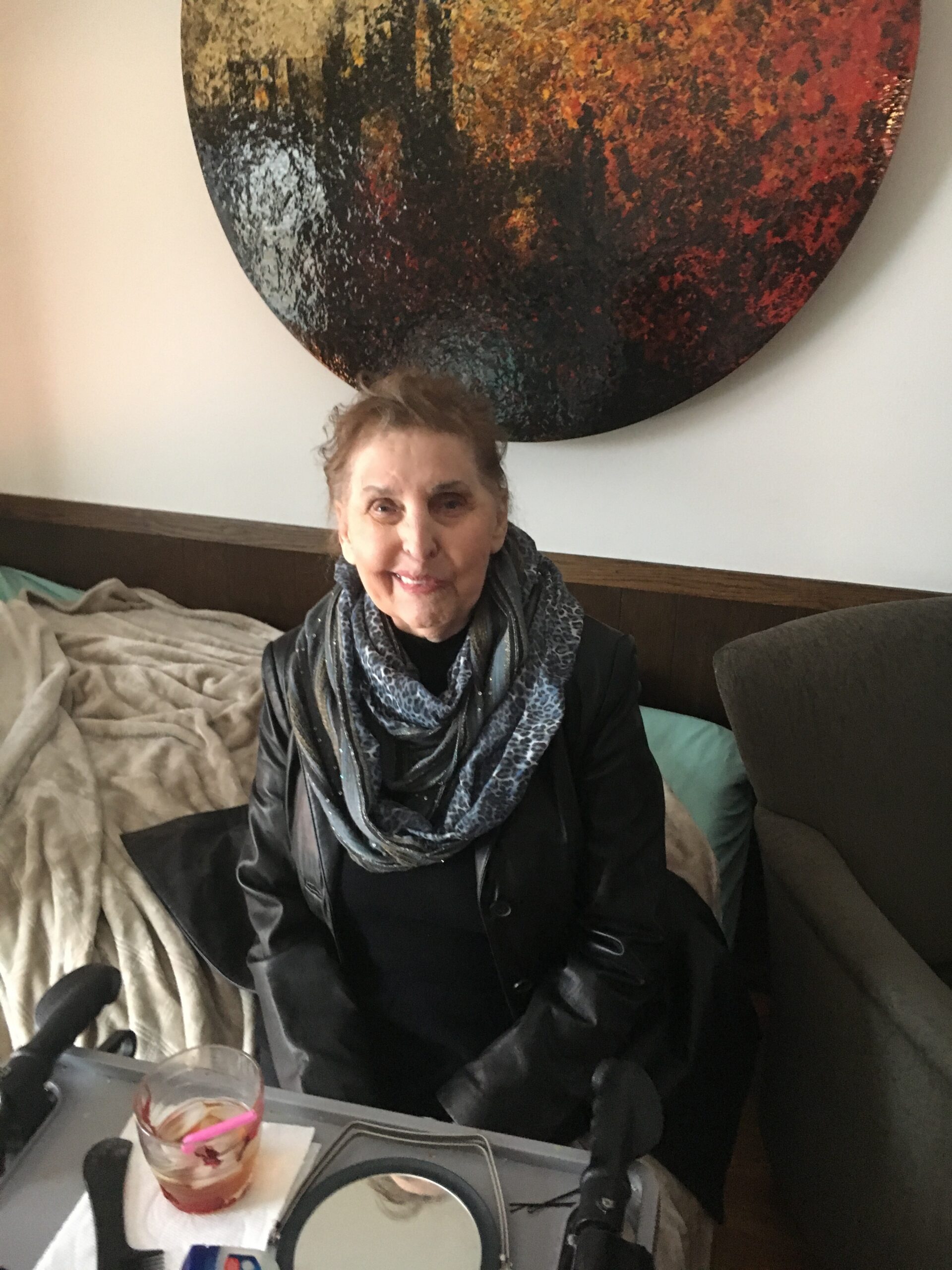 Smiling older woman in a black jacket and scarf sits on a couch with a large round abstract painting on the wall behind her.