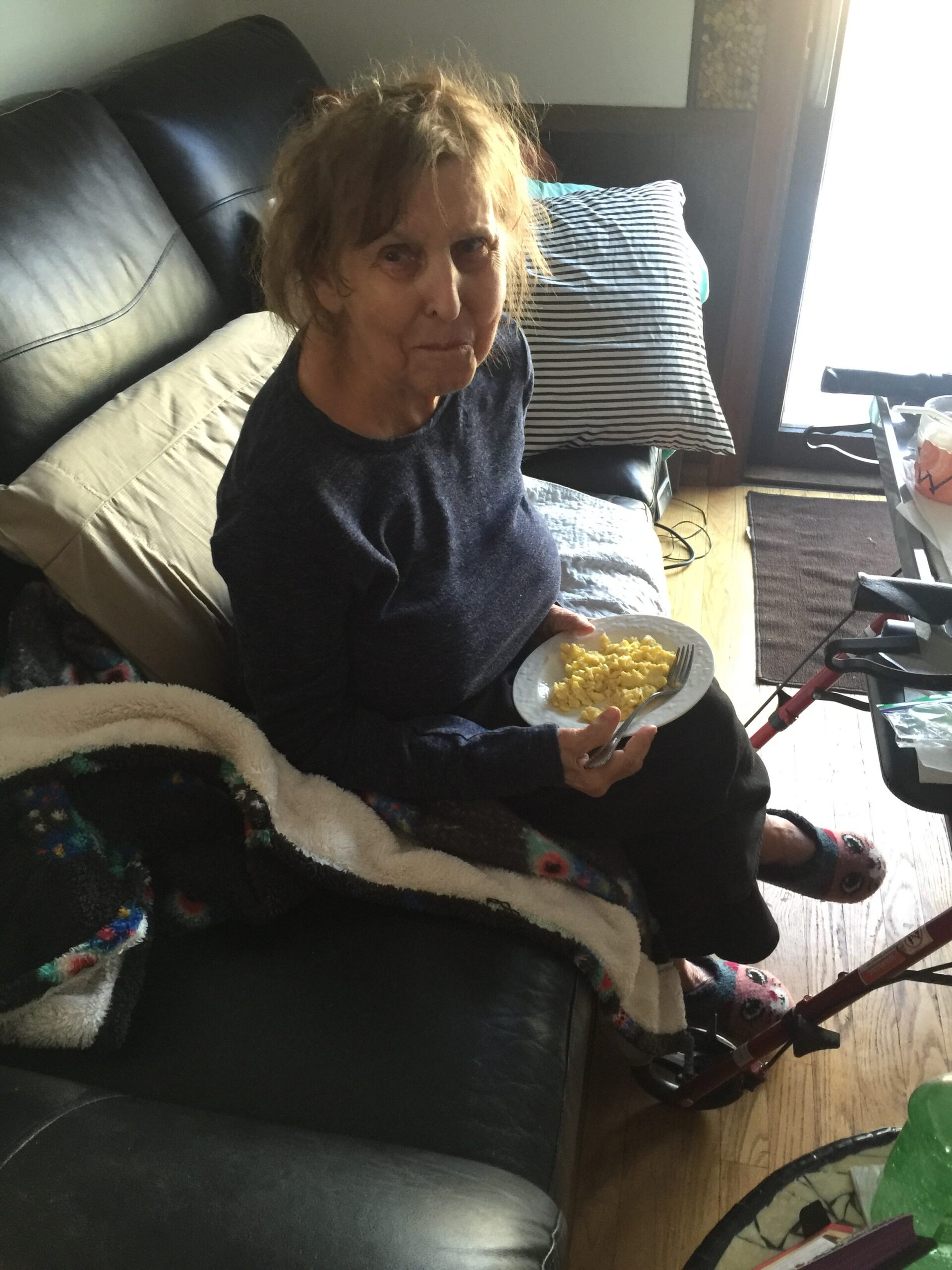 Older woman sits on a black leather couch eating scrambled eggs from a plate. A striped cushion and a blanket are nearby, with a walker visible by the window.