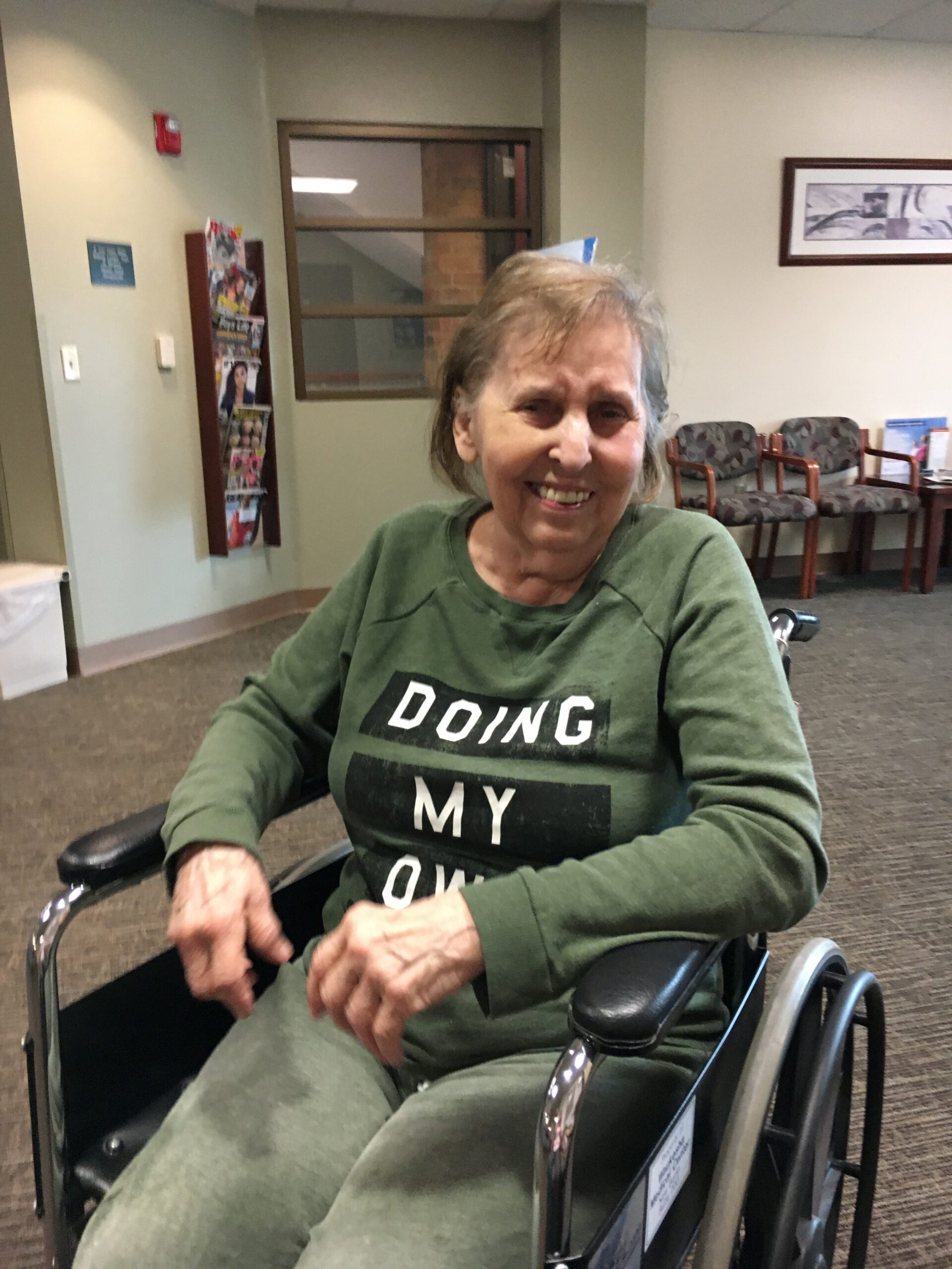Smiling elderly woman in a green shirt seated in a wheelchair in a waiting room. The shirt reads 'DOING MY OWN'.