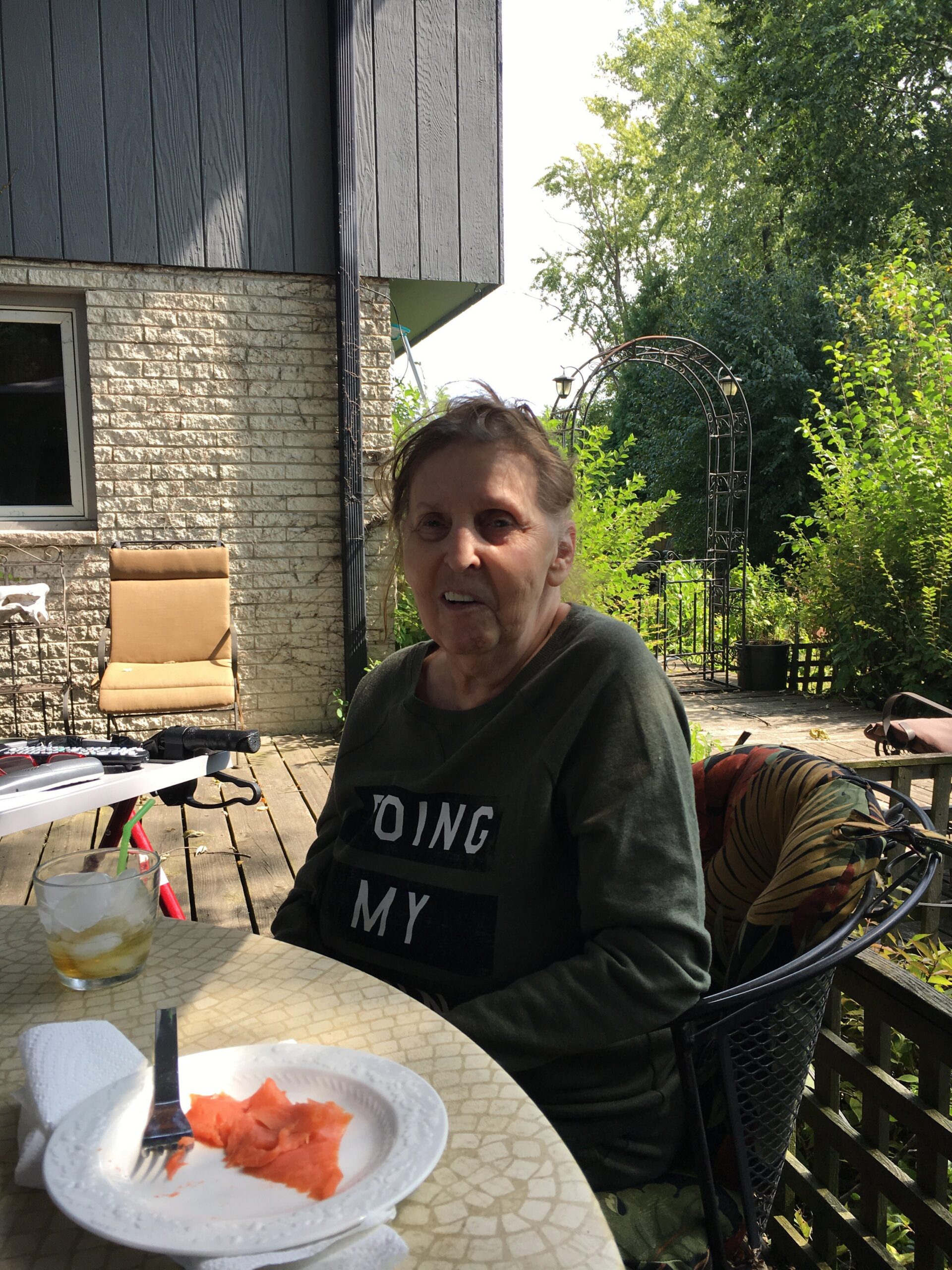 Older woman in a dark sweater sits at a patio table on a wooden deck, with a plate of watermelon and a glass in front, in a garden setting.