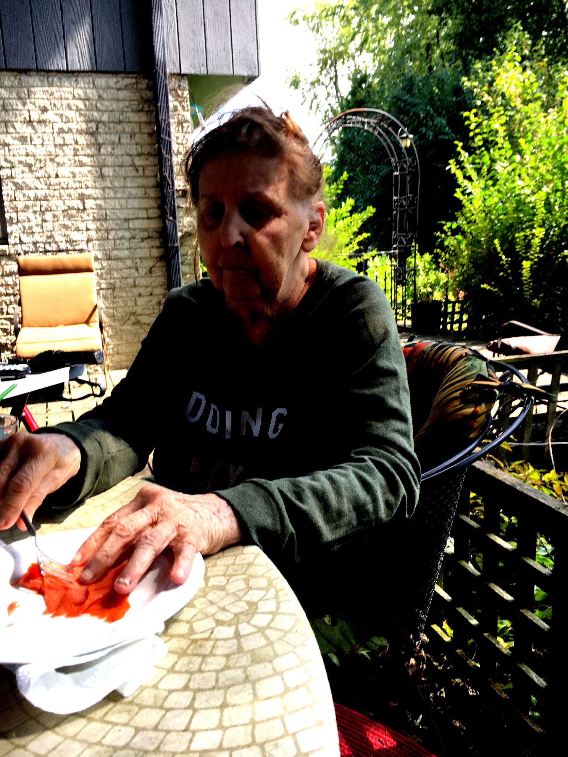 Older person seated at a sunlit outdoor table, cutting or handling slices of watermelon on a plate.