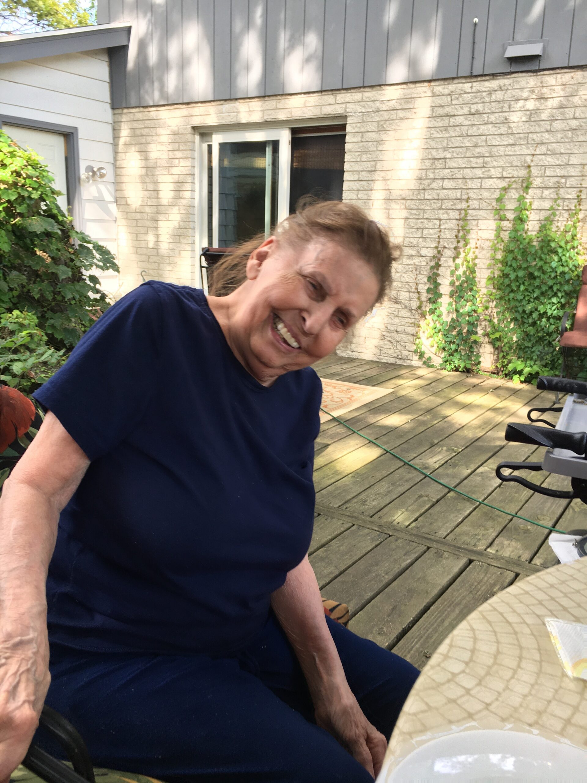 Smiling elderly woman in a dark navy shirt sits on a wooden deck outdoors, with a brick house and green vines in the background.