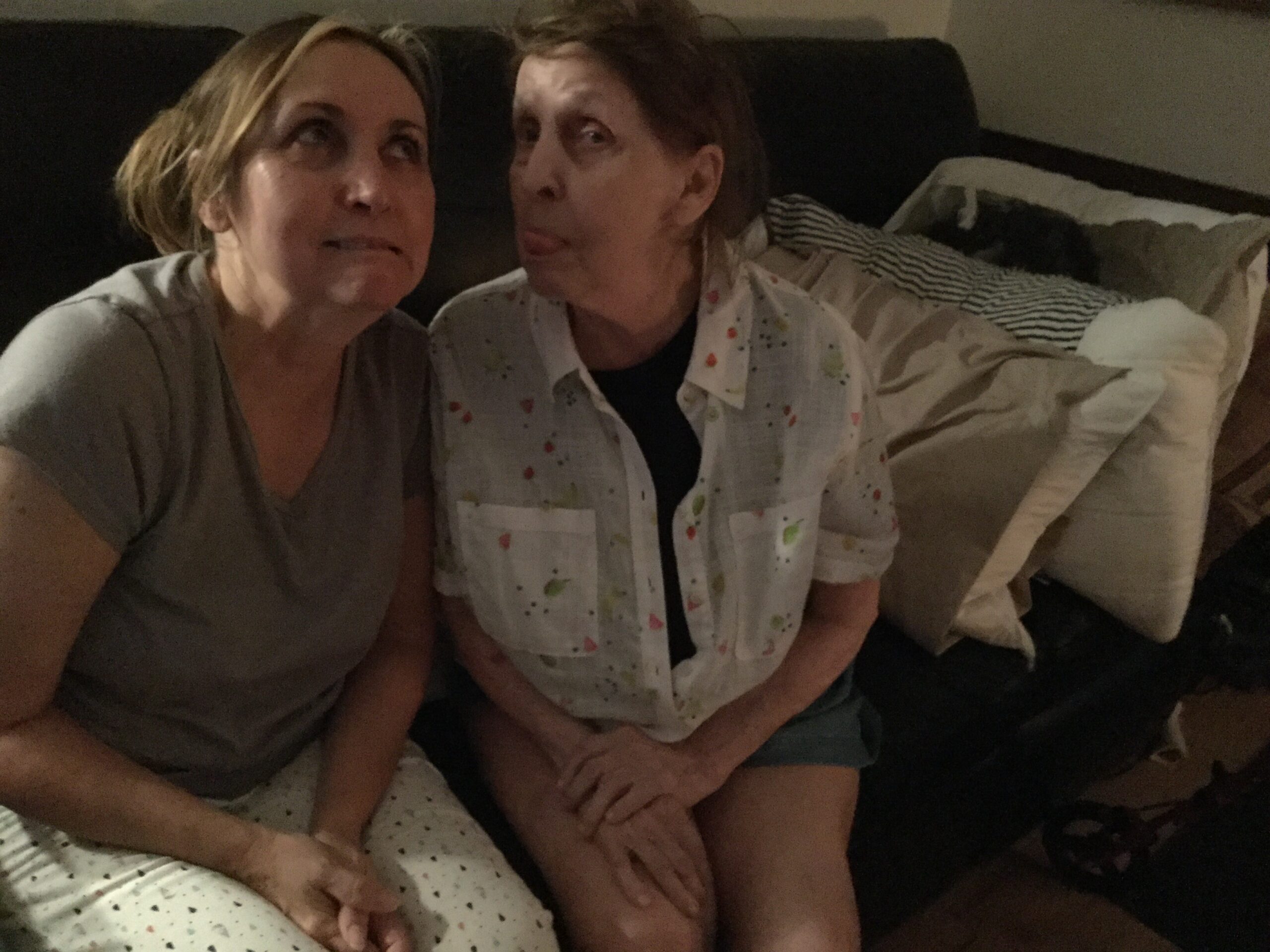 Two women sit close on a sofa in a dim room, facing each other and smiling with playful expressions.