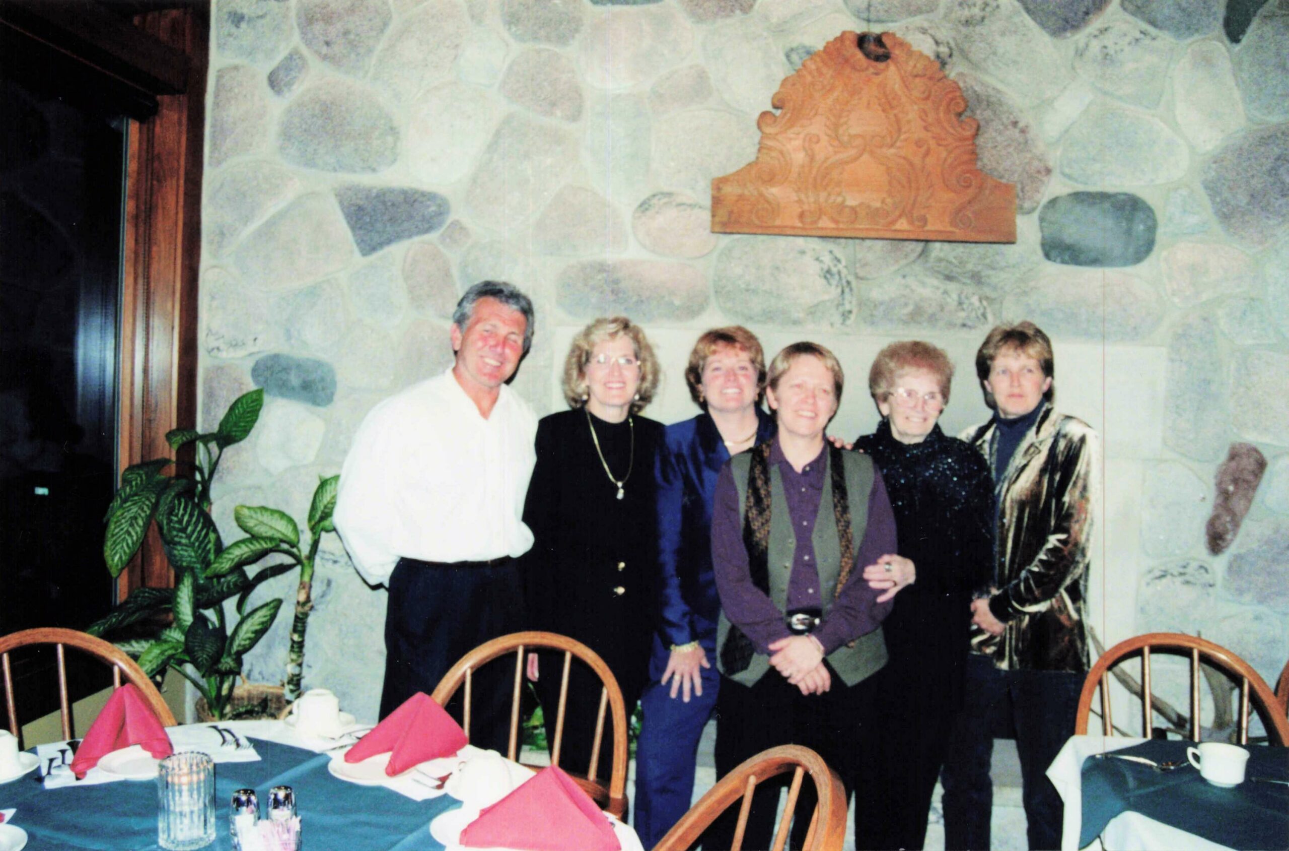 Six adults pose together for a group photo in a stone-wall dining room with a carved wood plaque above them, near a table set with red napkins.