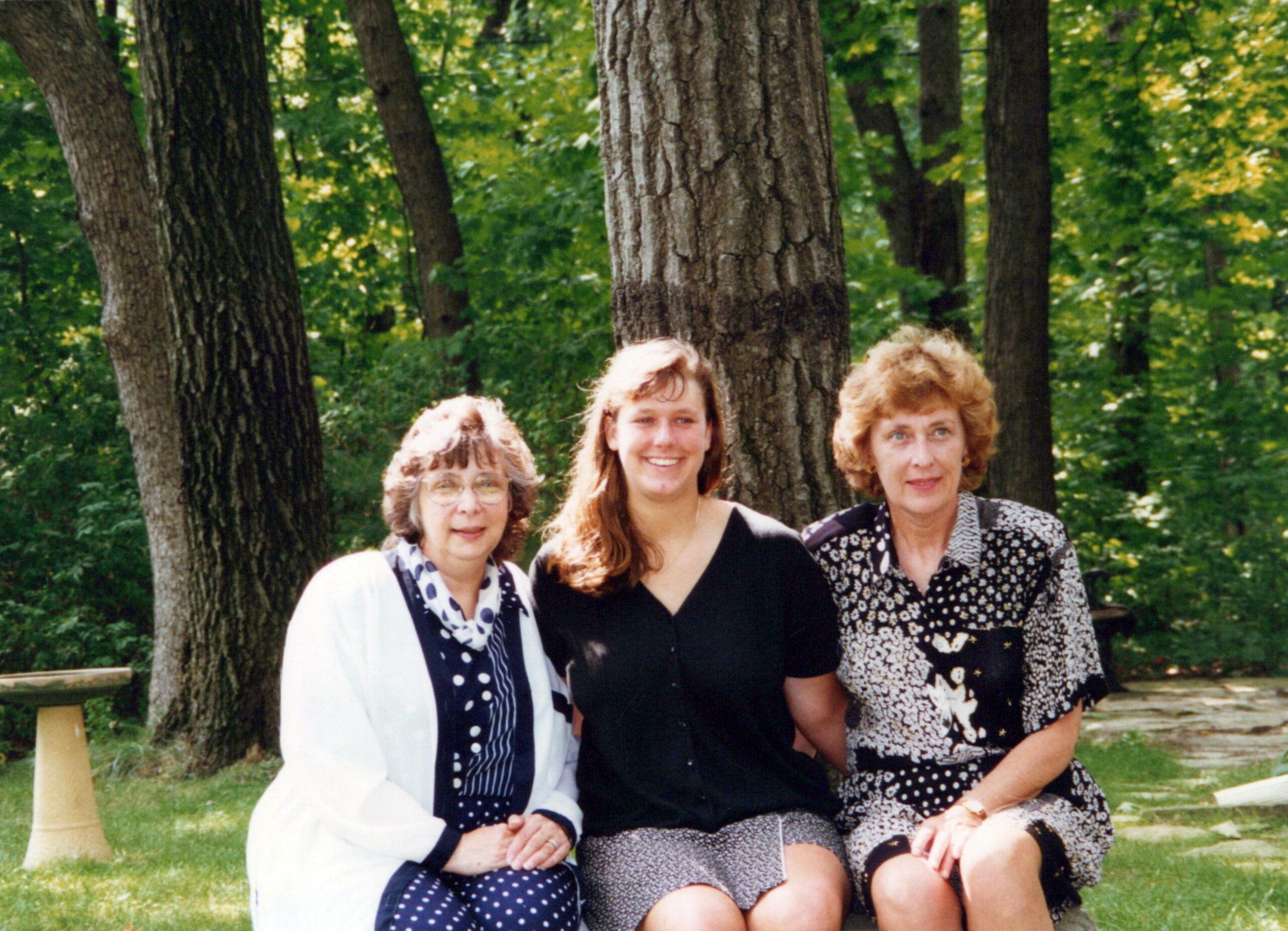 Four women sitting close together outdoors in a wooded park, posing for a photo with trees in the background, smiling at the camera