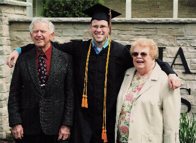 College graduate in cap and gown posing with an older man and woman outdoors, all smiling with arms around each other.