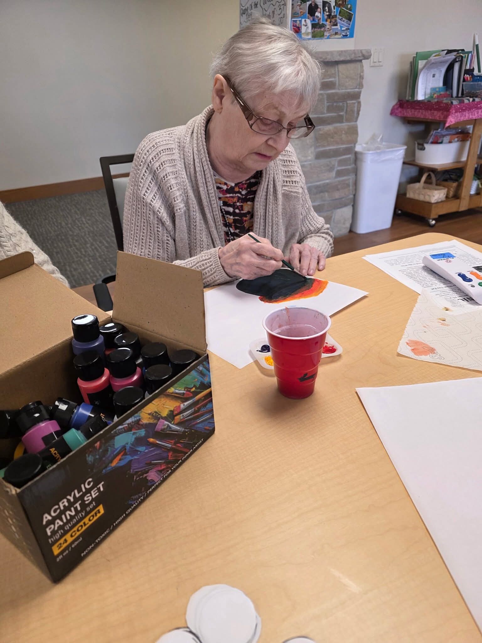 Older woman painting with a brush at a table, acrylic paints box open nearby and a red cup on the desk.