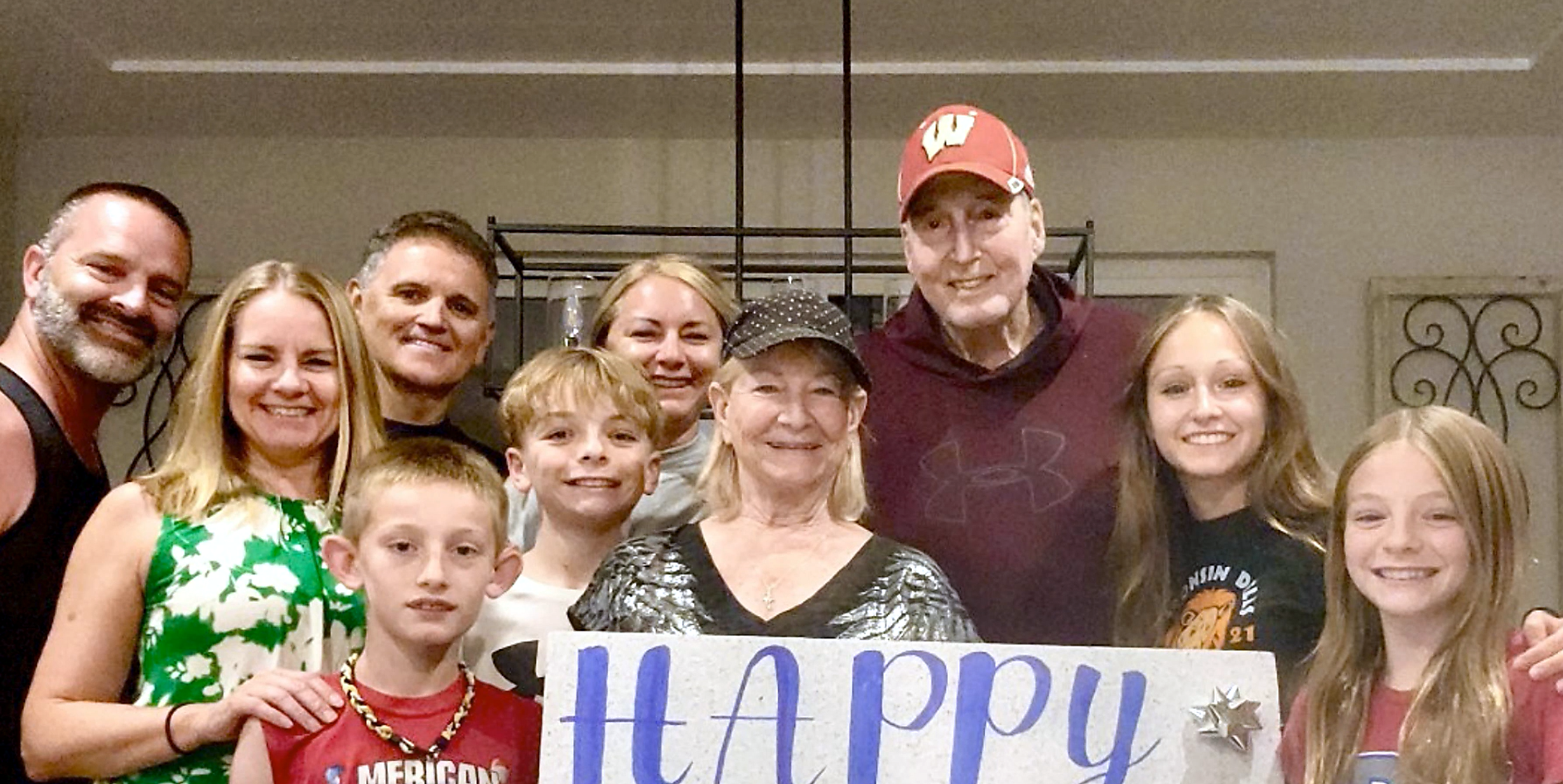 Group of smiling people of various ages posing indoors with a large 'Happy' sign for a celebration (family/friends photo).