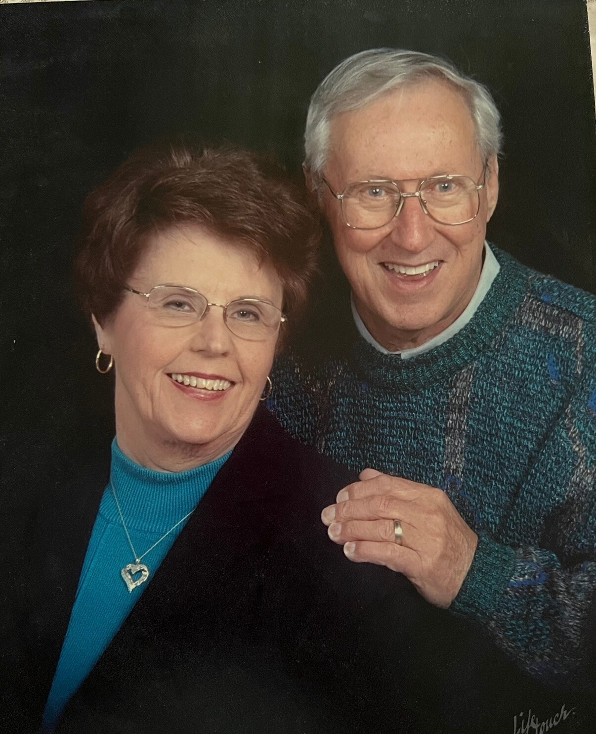 Happy elderly couple posing together and smiling at the camera; she wears a turquoise turtleneck with a heart pendant while he wears glasses and a green knit sweater.