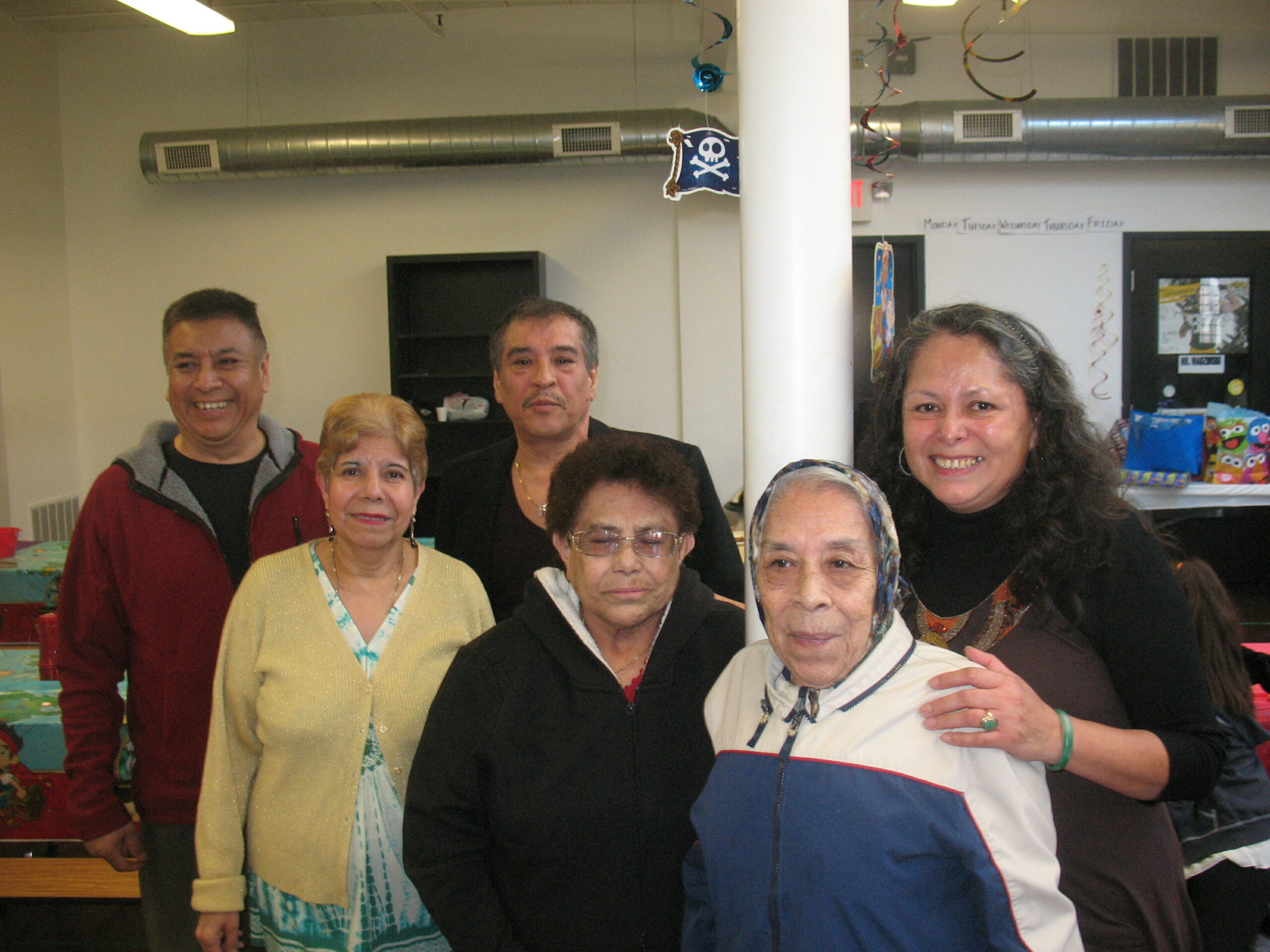 Group of smiling adults posing for a photo at an indoor celebration, with festive streamers and decorations in the background.