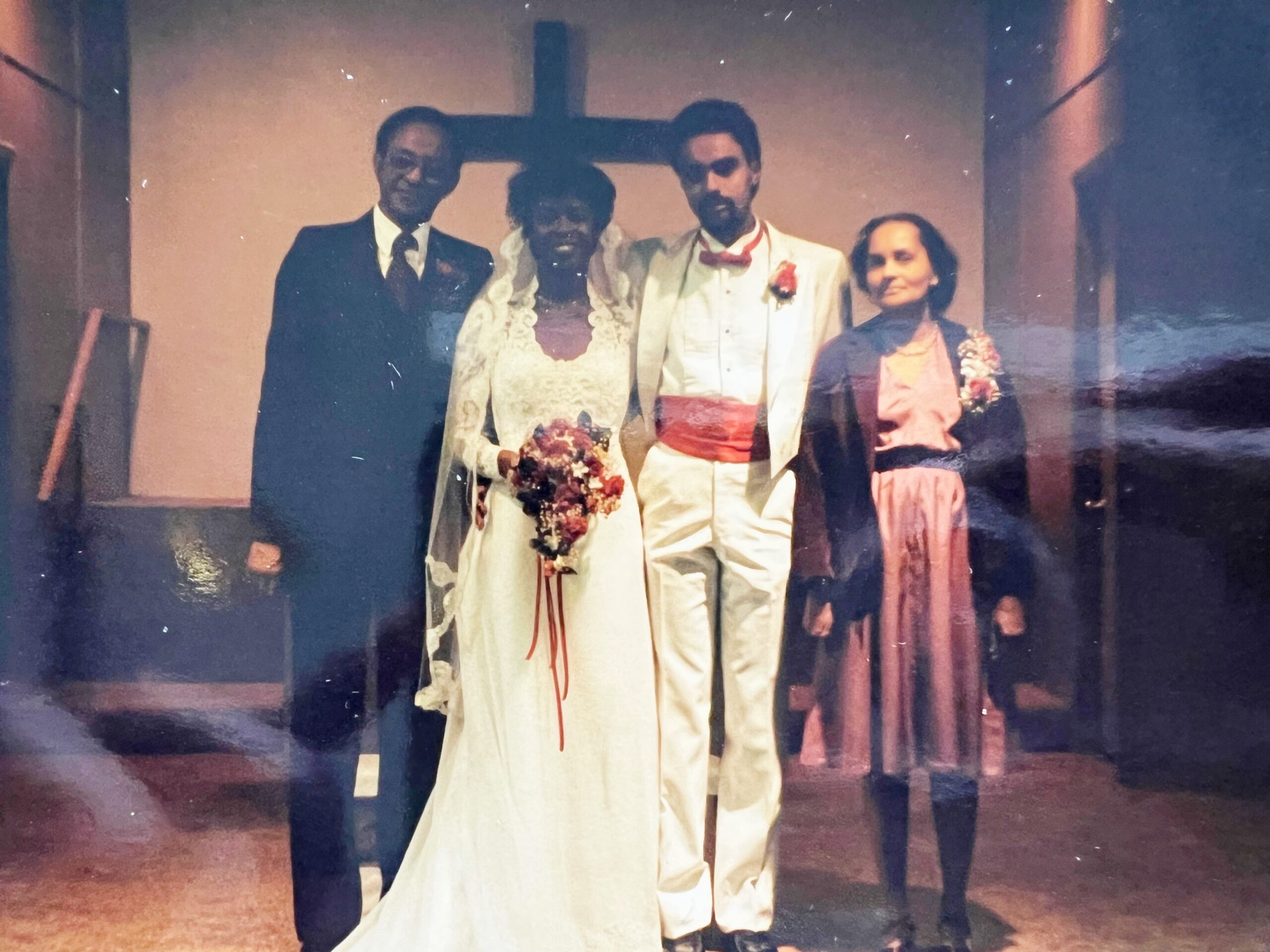 Group portrait of a wedding party: bride in lace gown and groom in white suit with red sash, flanked by a man in a dark suit and a woman in a pink dress, with a cross in the background.