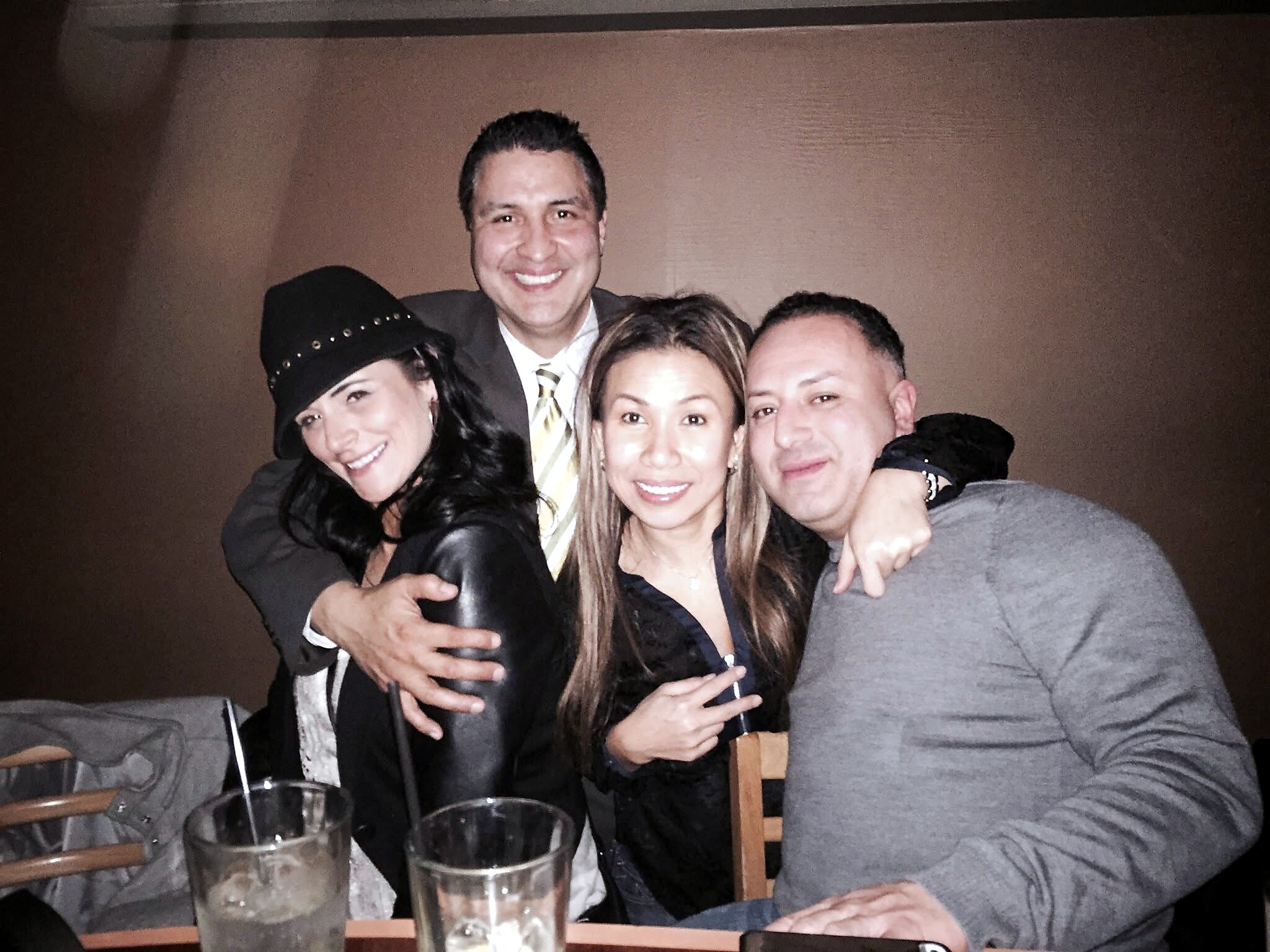 Group of four friends posing for a selfie in a dimly lit setting, smiling with drinks on the table in front of them, arms around each other.