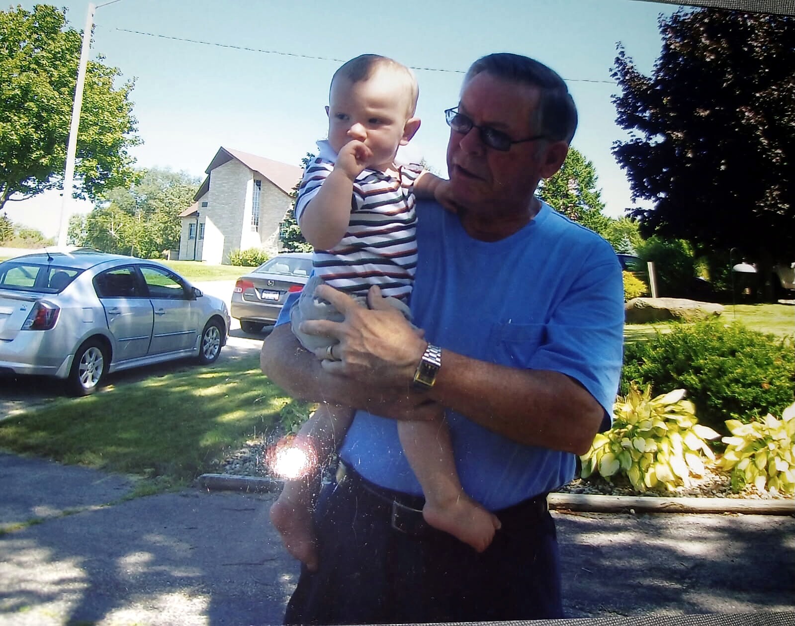 Man in a blue shirt holding a baby in a striped outfit, outdoors in a sunny suburban yard with parked cars and trees.