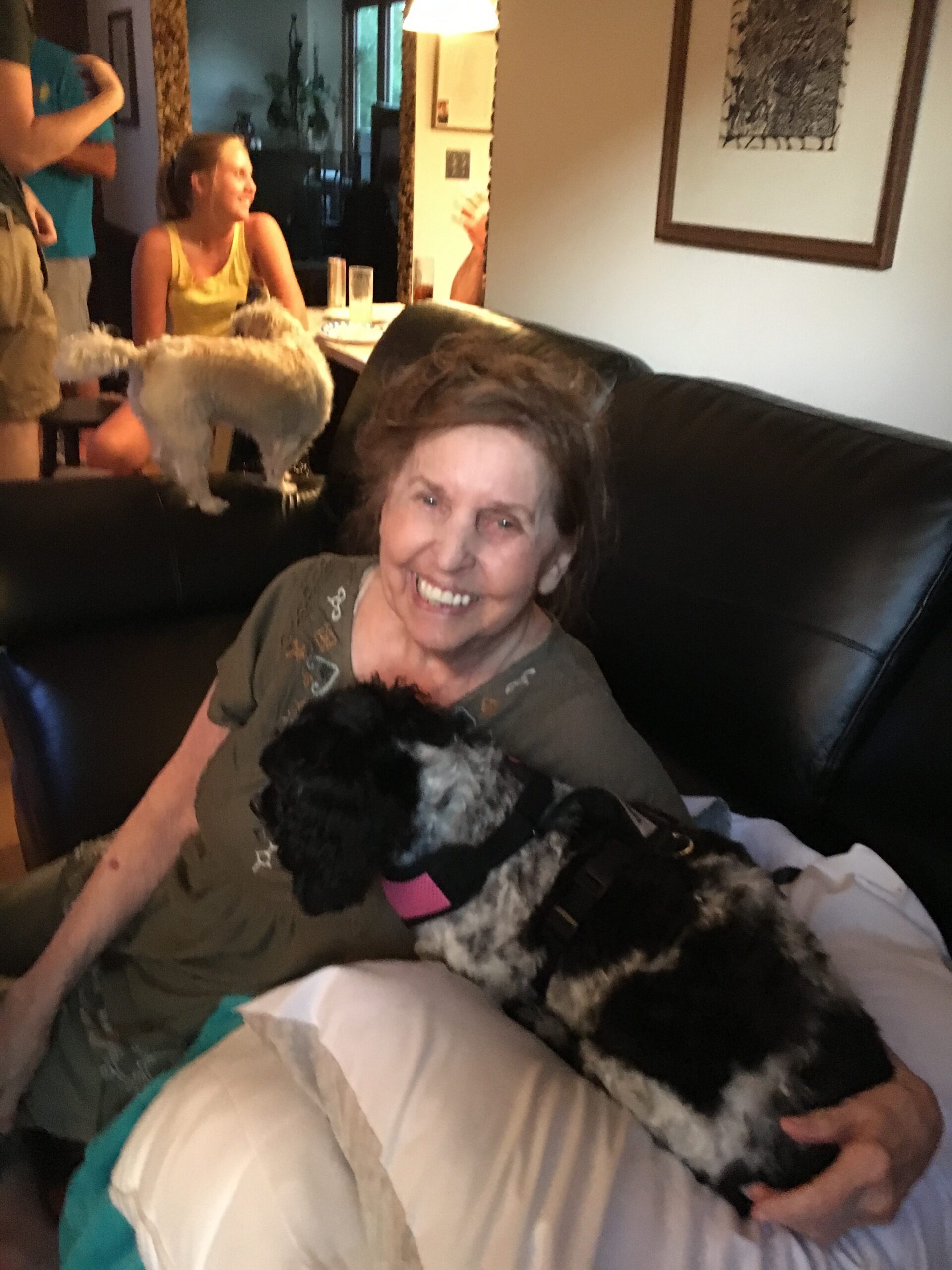 Smiling elderly woman on a dark leather couch holding a black-and-white dog, with a white fluffy dog nearby and people in the background in a living room.