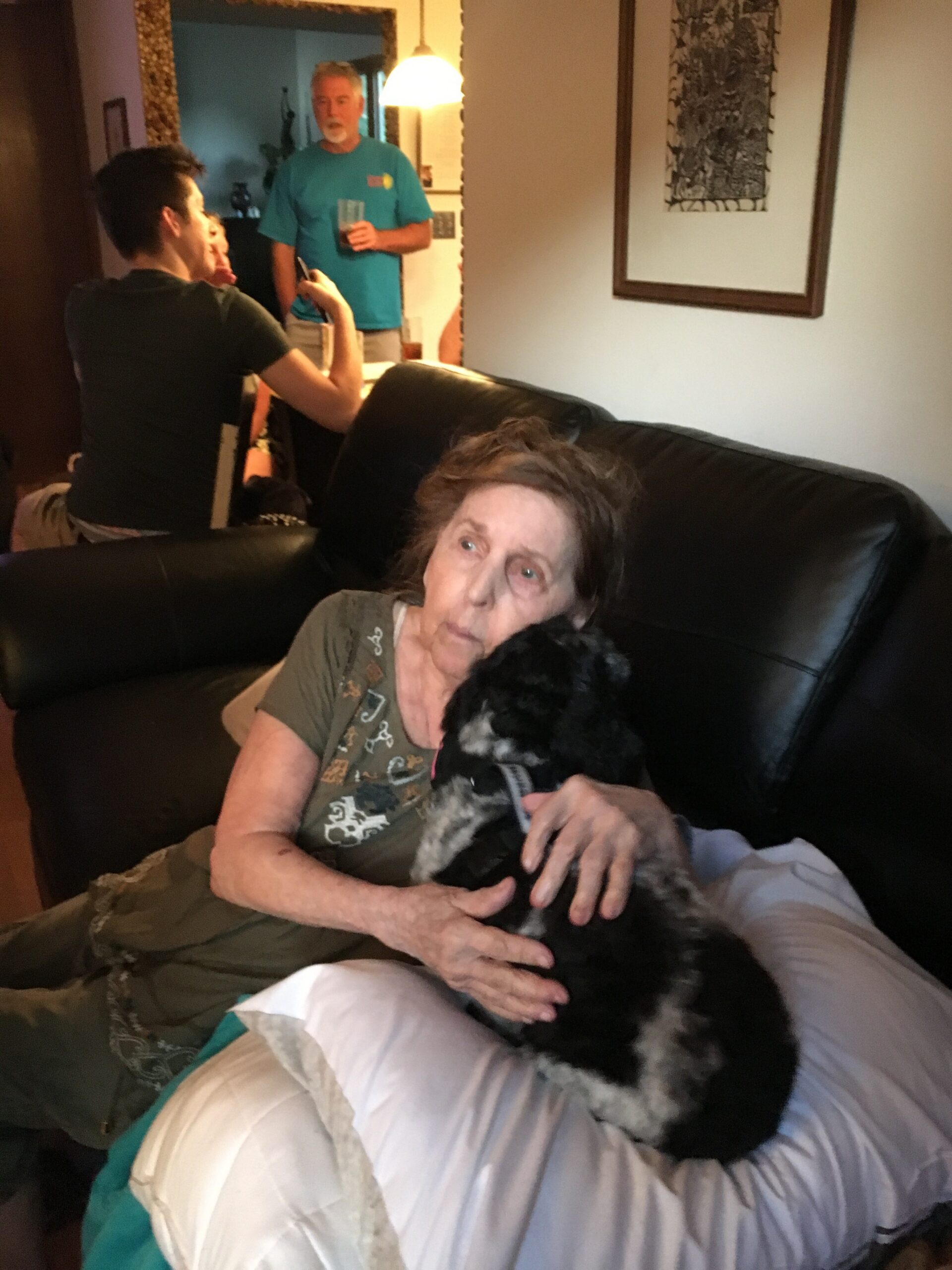 Woman sitting on a couch cuddling a black fluffy dog as a man in a teal shirt stands in a doorway holding a drink in the background.