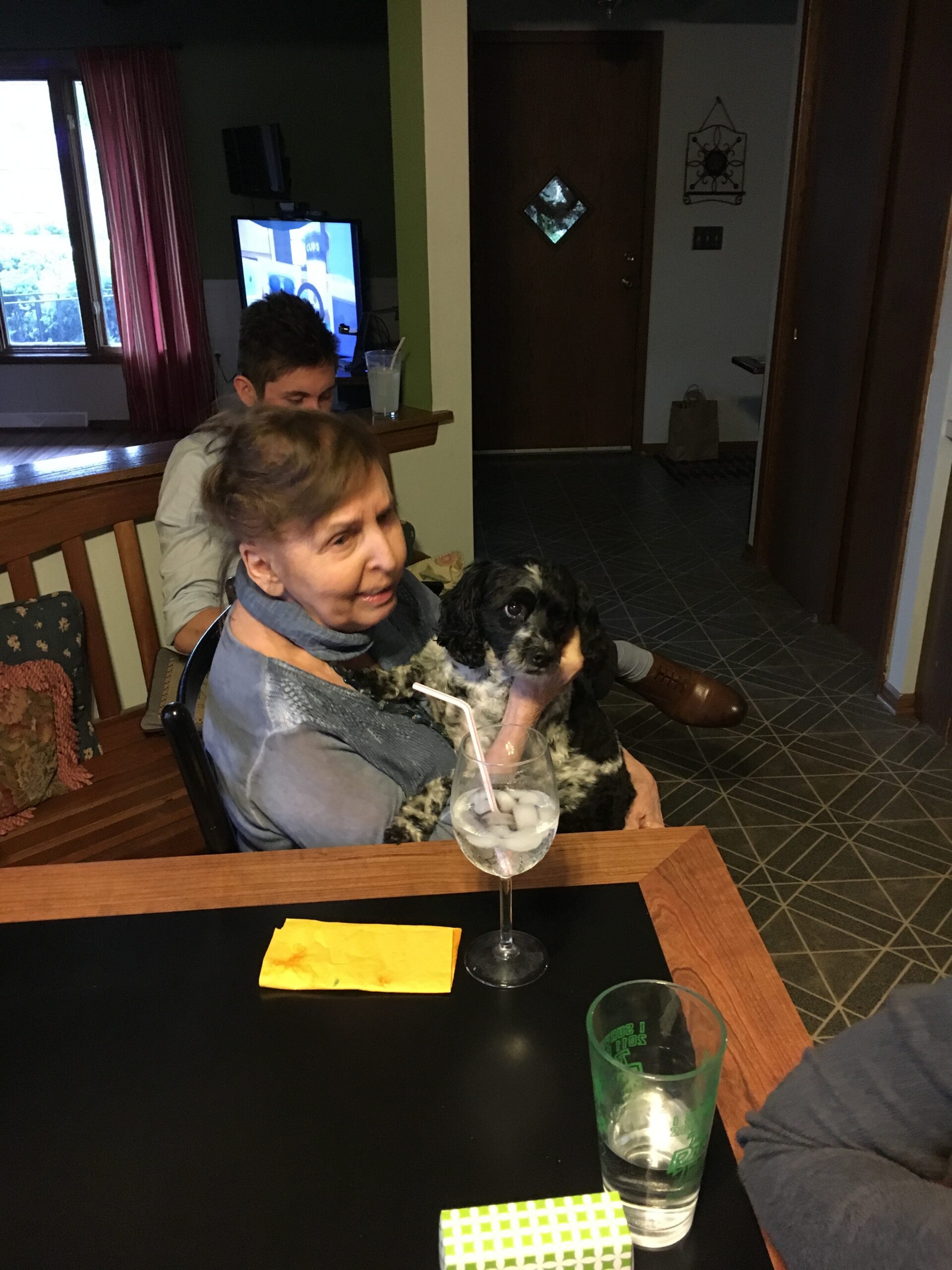 Older woman holding a black-and-white dog on her lap at a dining table, with a man behind her and a TV in the background.