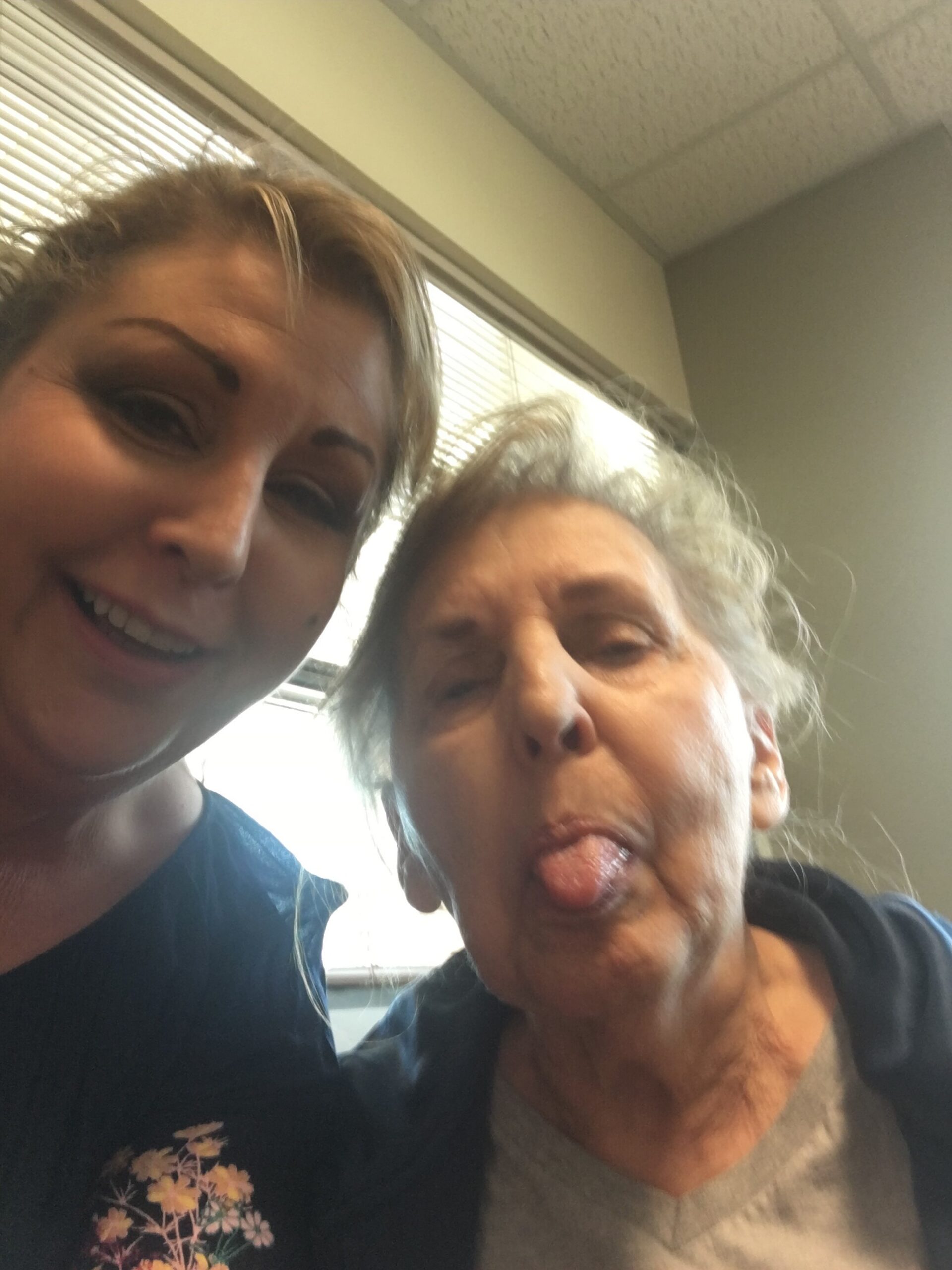Two women share a playful selfie indoors, one smiling at the camera and the other sticking out her tongue near a window with blinds behind them.