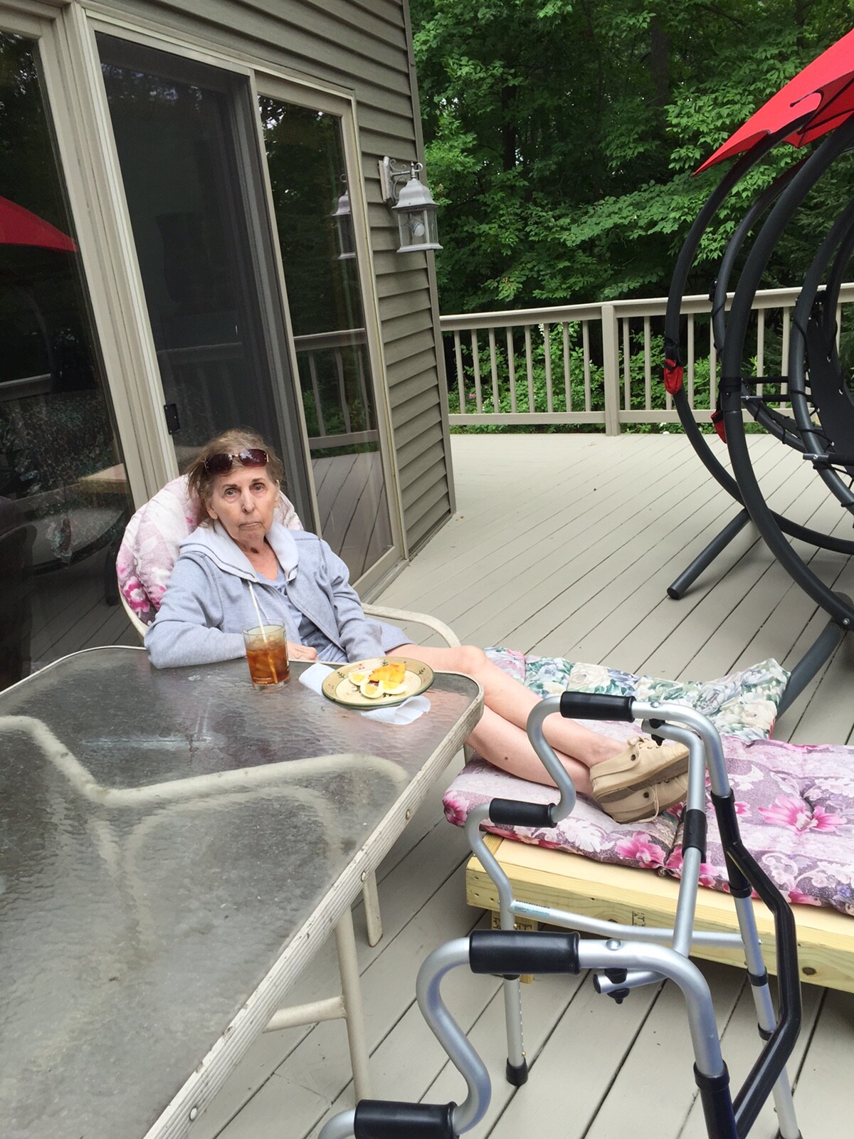 Older woman sitting in a cushioned chair on a wooden deck, with a plate of fruit and iced tea, and a walker in the foreground.