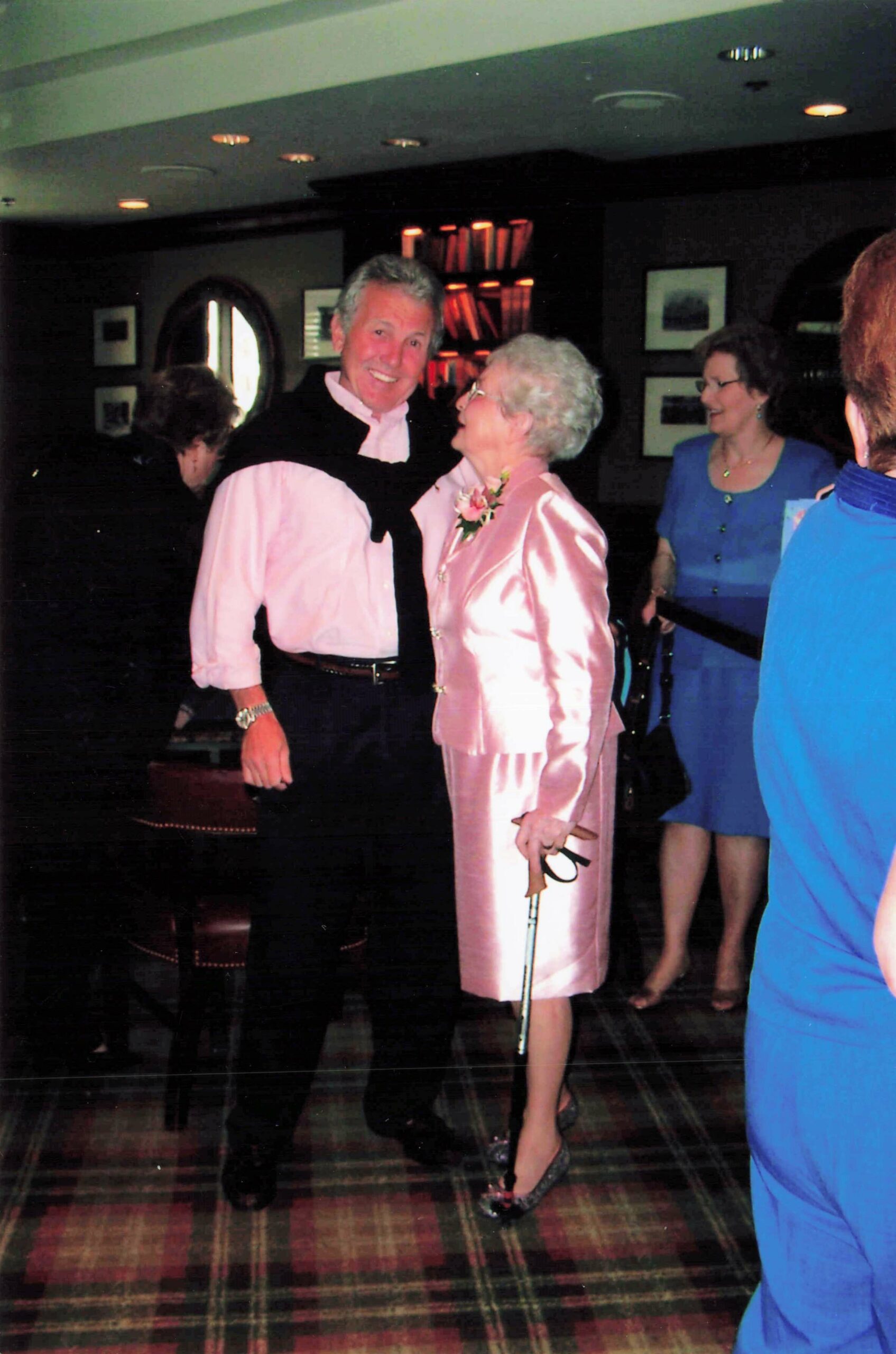 Smiling elderly couple posing at a formal party: man in pink shirt with sweater draped over shoulders, woman in pink satin suit with corsage, others in background