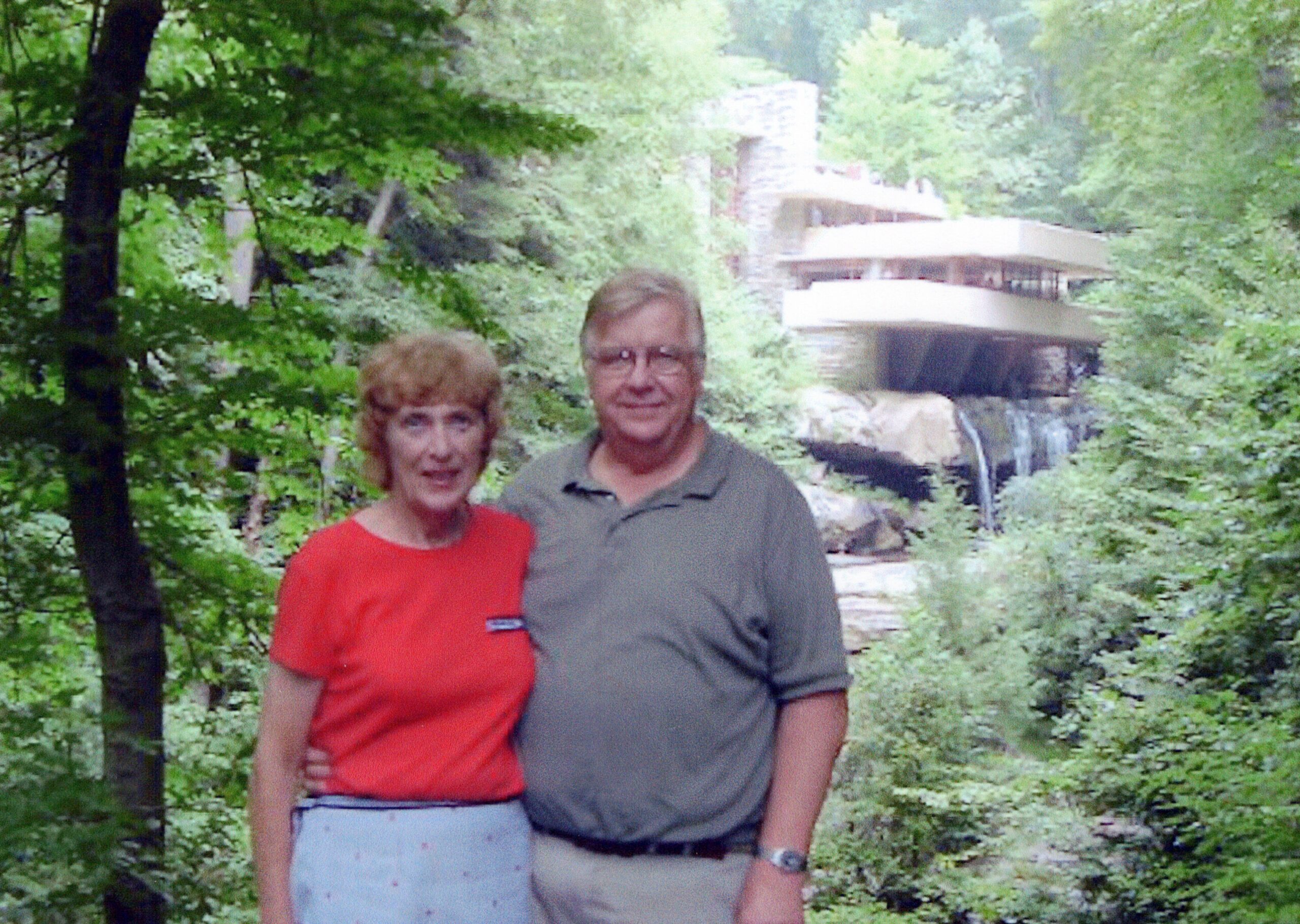 An older couple stands together in a green forest with a small waterfall and a modern building visible in the background.