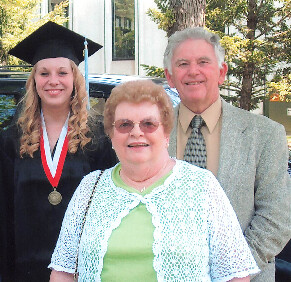 Young graduate in cap and gown posing outdoors with her mother and father.