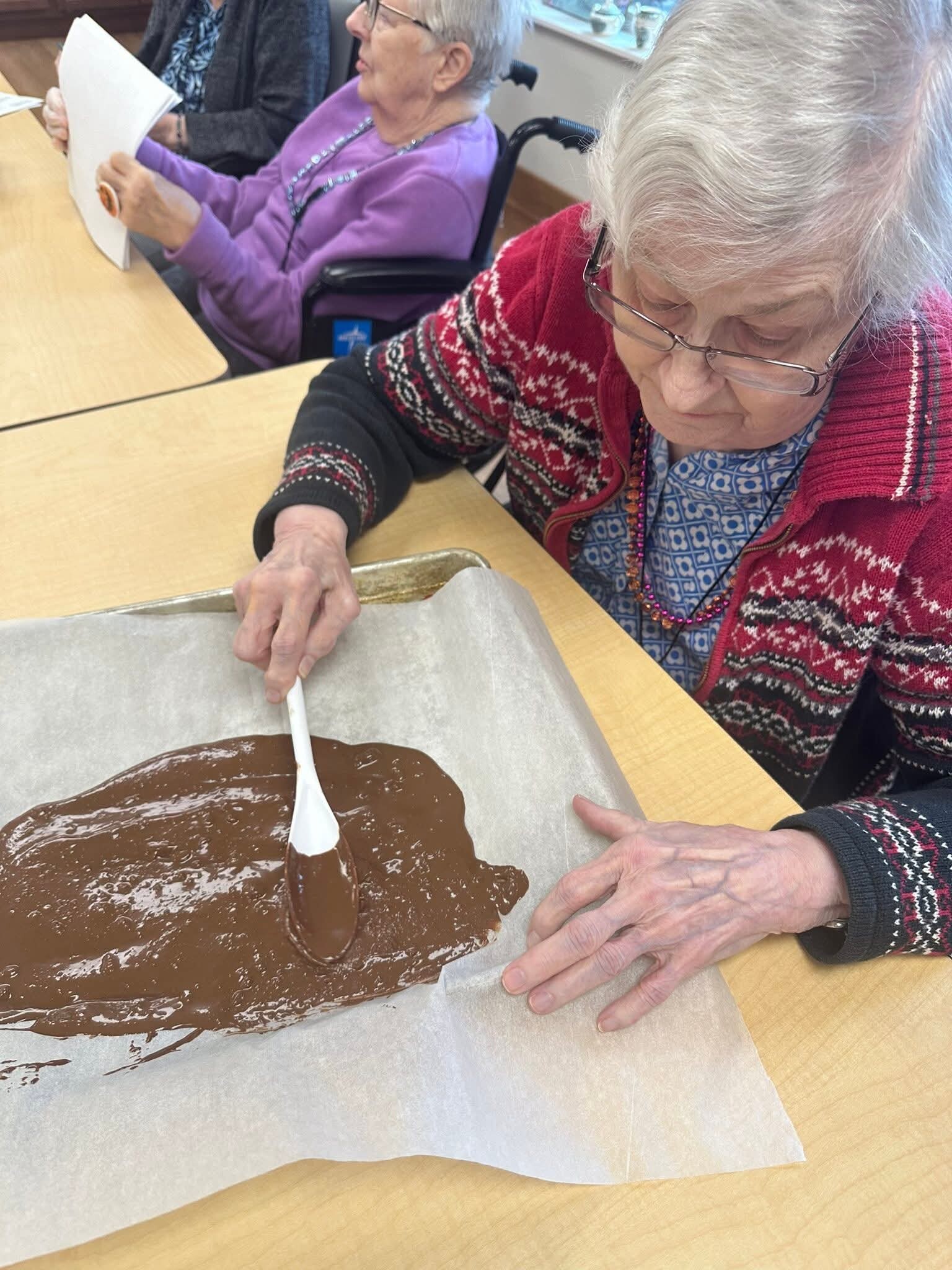 Two elderly women sit at a table; one in a red patterned cardigan stirs melted chocolate on parchment with a white spatula, while the other in a purple top looks at papers.
