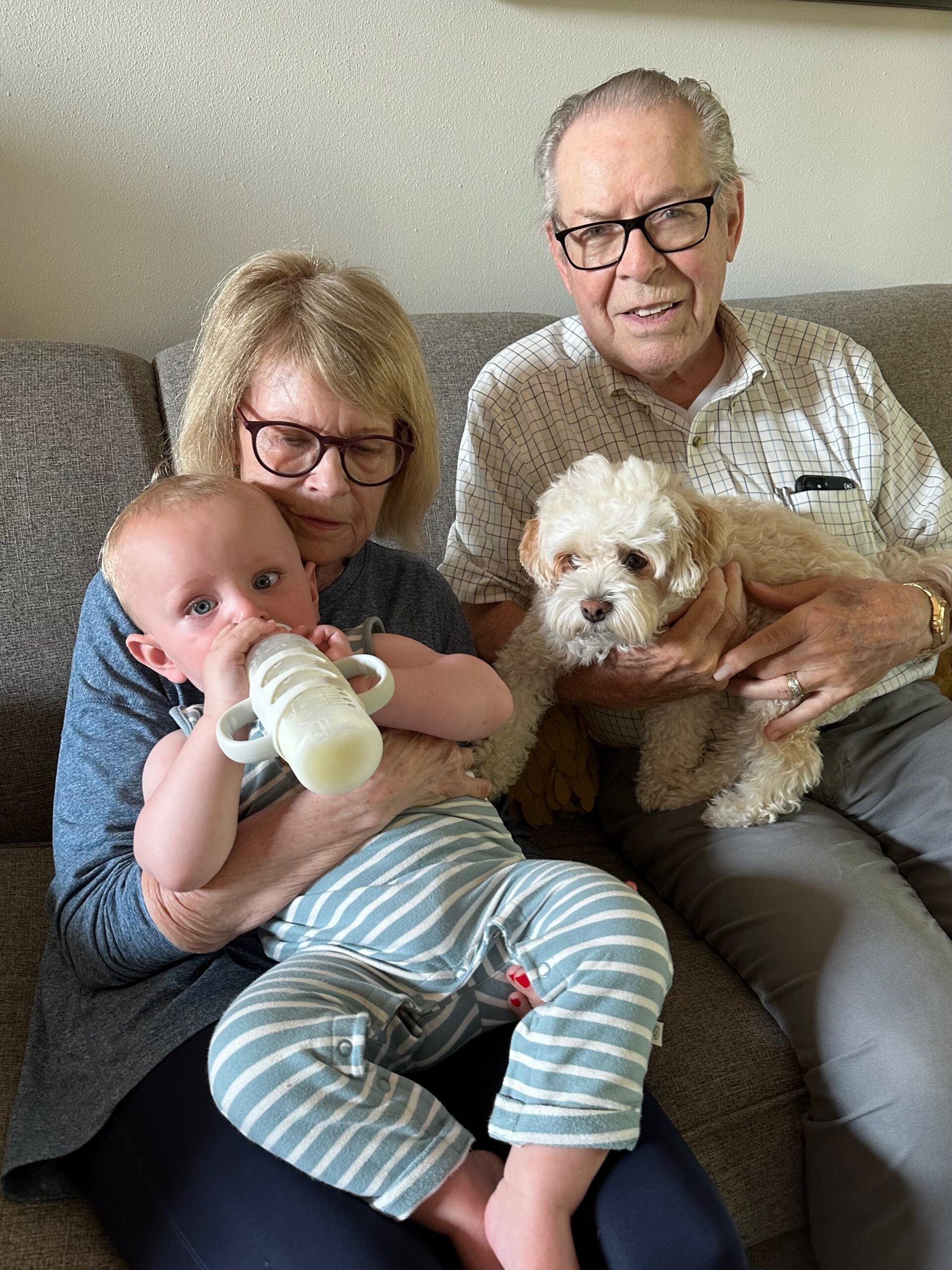 Grandmother on a sofa holding a baby bottle while a small fluffy dog sits with grandfather nearby.