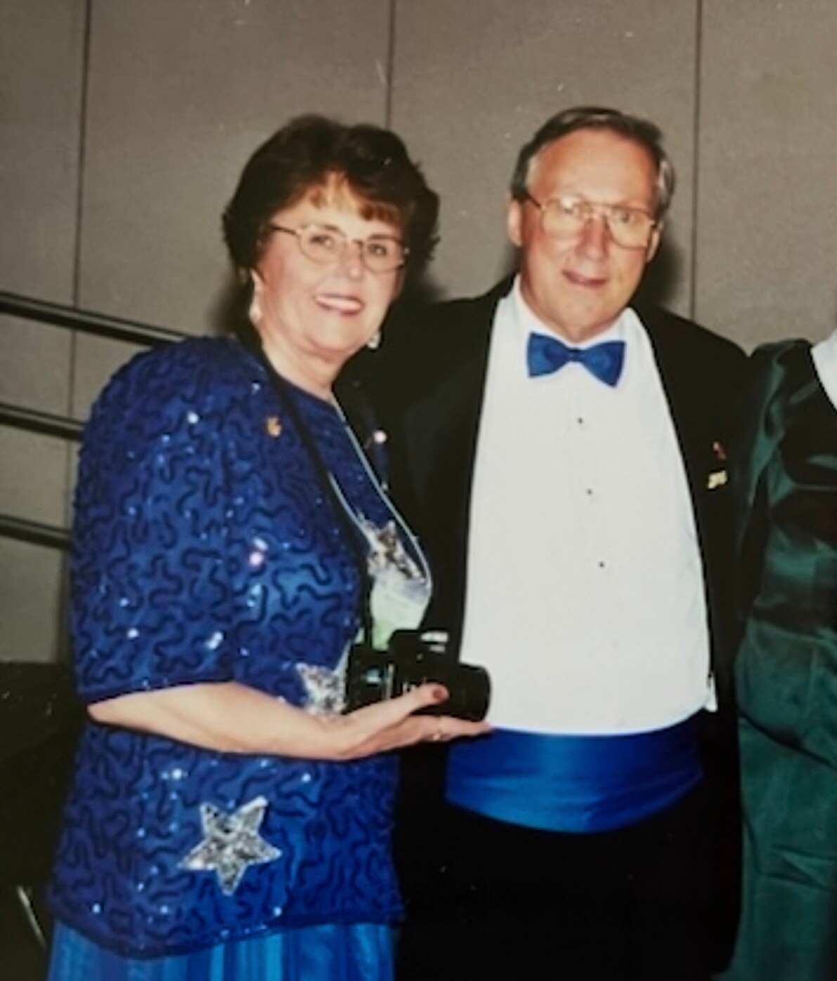 Smiling elderly couple at a formal event: woman in a blue sequined dress with a star pin, man in a black tuxedo with a blue bow tie.