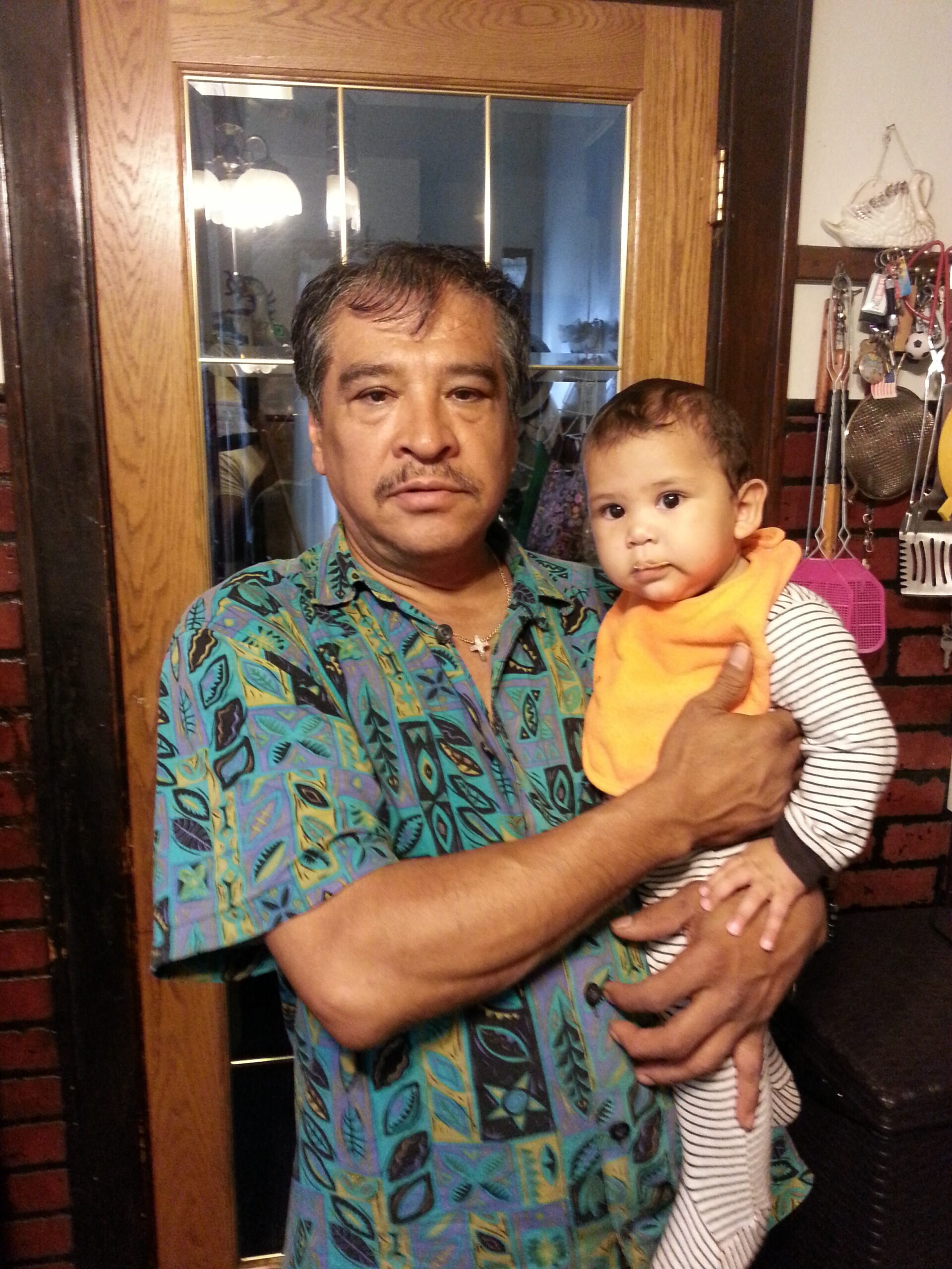 Man in a teal patterned shirt holding a baby in an orange bib, standing in a doorway indoors.