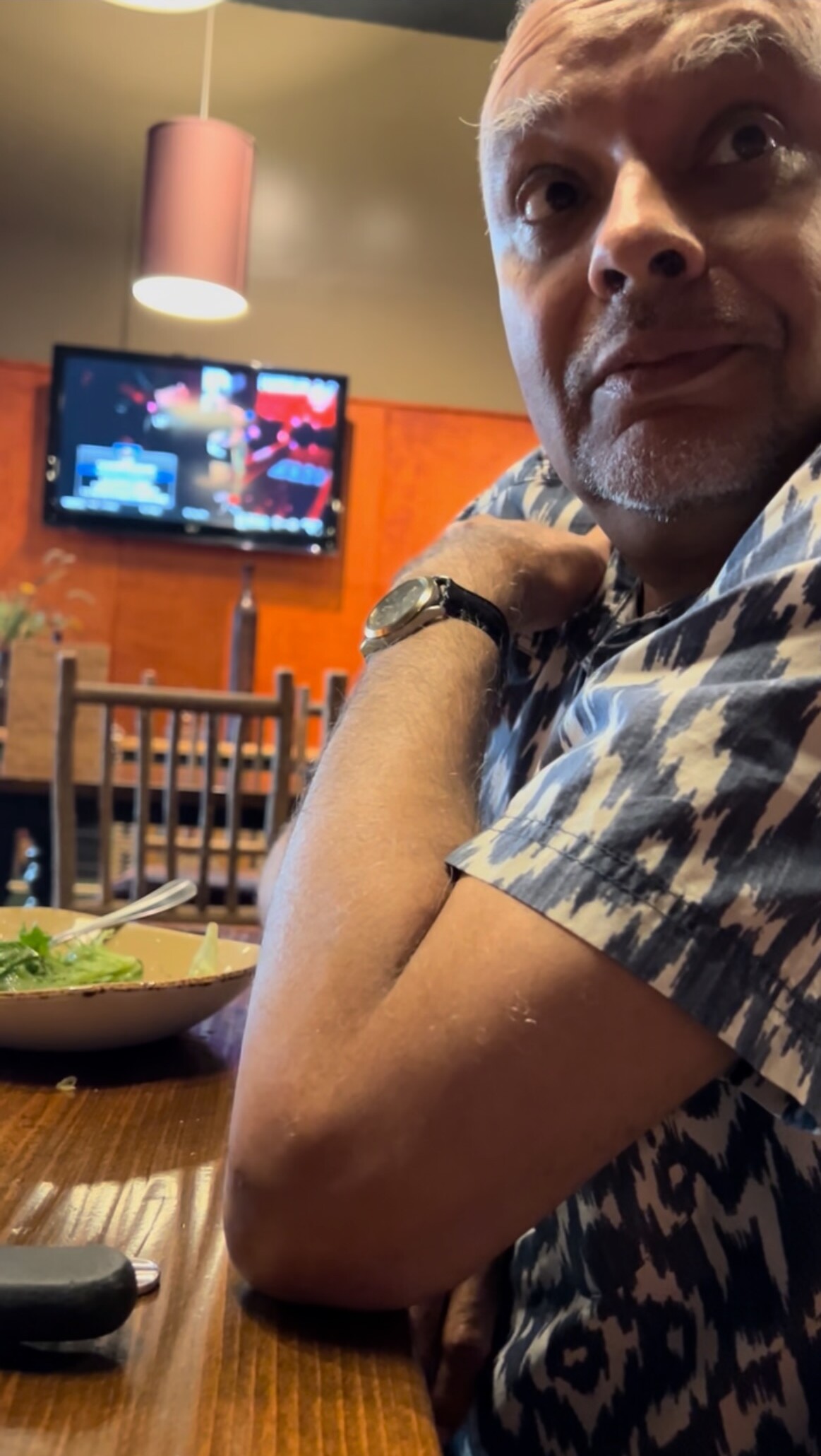 Man wearing a blue‑white patterned shirt sits at a wooden restaurant table, resting his arm; salad bowl in foreground and a TV on the wall behind him.