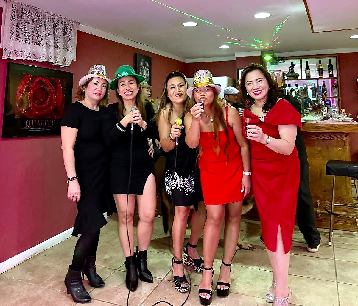 A group of five women posing and singing into microphones at a festive party in a bar, wearing colorful hats and dresses.