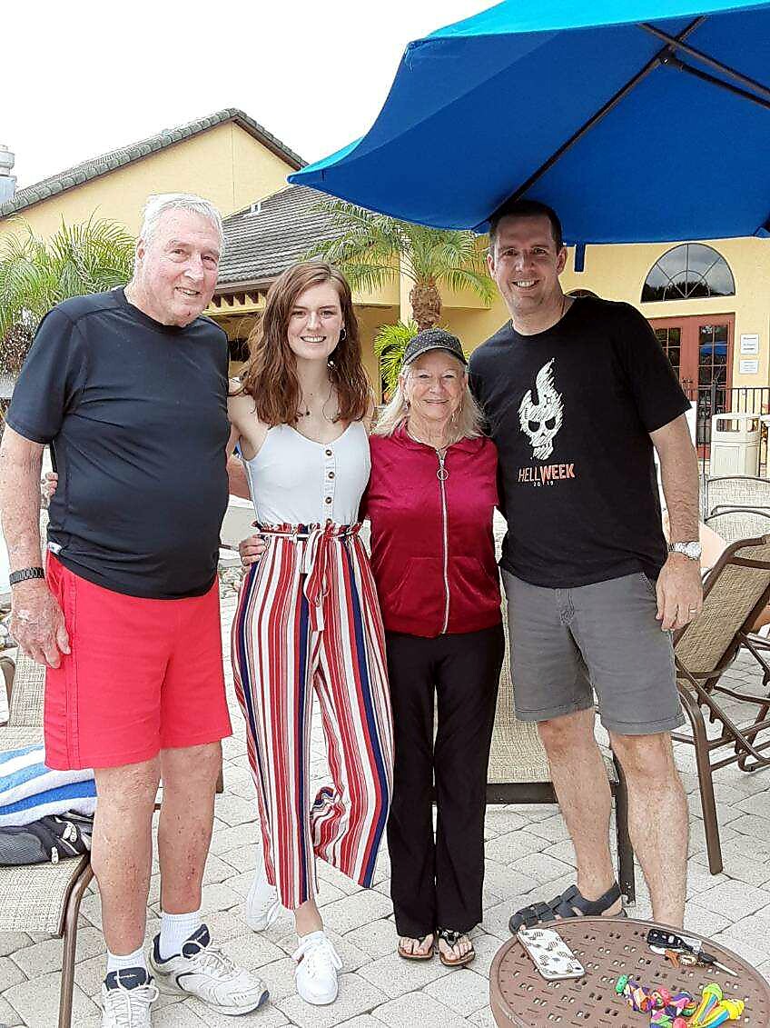 Four people posing outdoors under a blue umbrella, smiling at the camera. Left to right: man in black shirt and red shorts, woman in striped pants, older woman in red jacket, man in black t‑shirt.