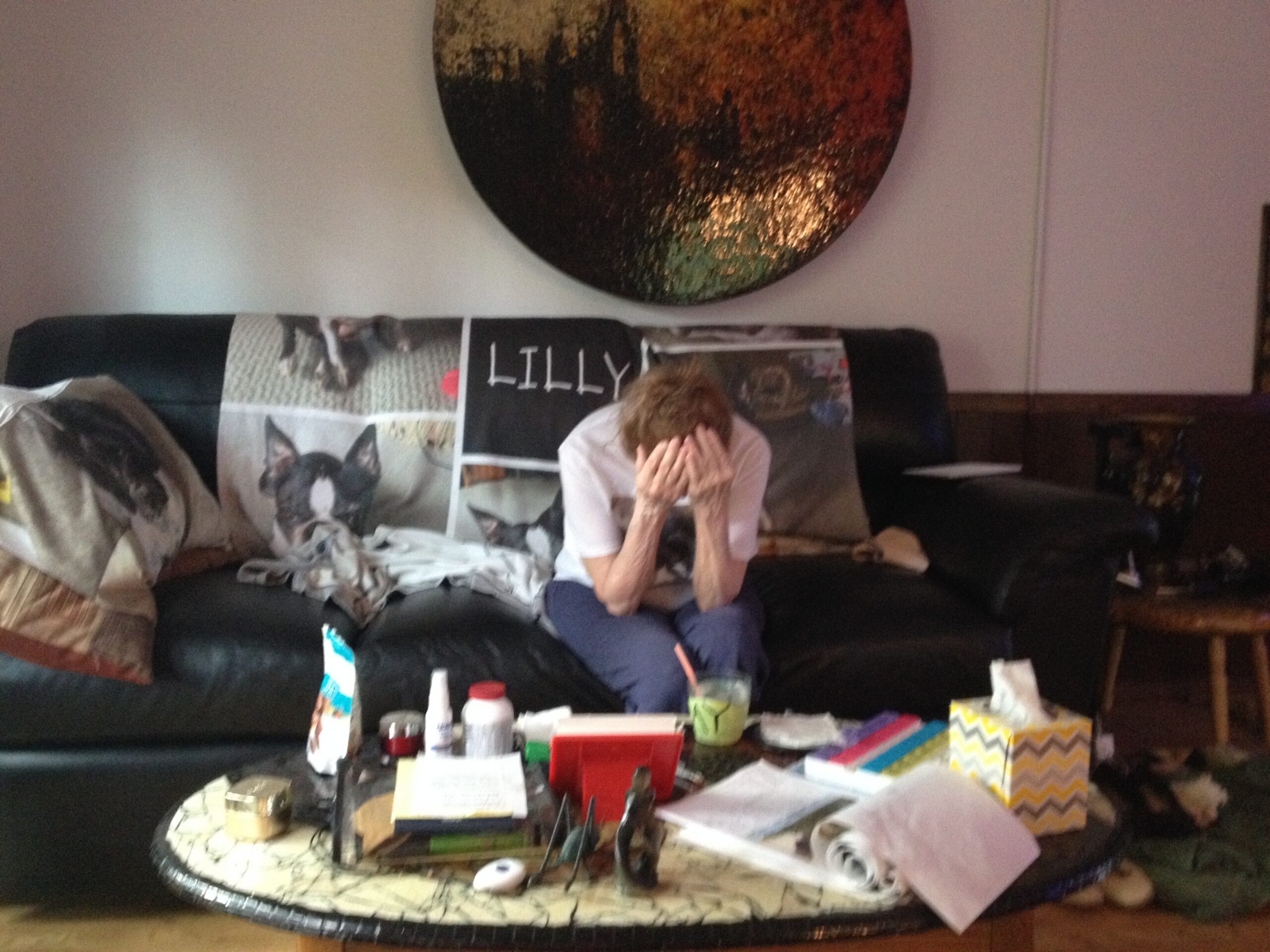 Person sitting on a black sofa with hands over their face in a cluttered living room, surrounded by posters, crafts, and scattered papers on a coffee table under a large circular wall art piece