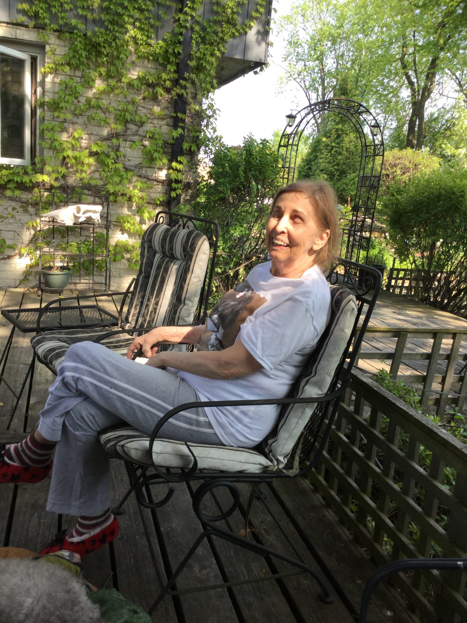 Older woman sits smiling on a striped patio chair on a wooden deck, ivy-covered house wall in a sunny garden behind her.