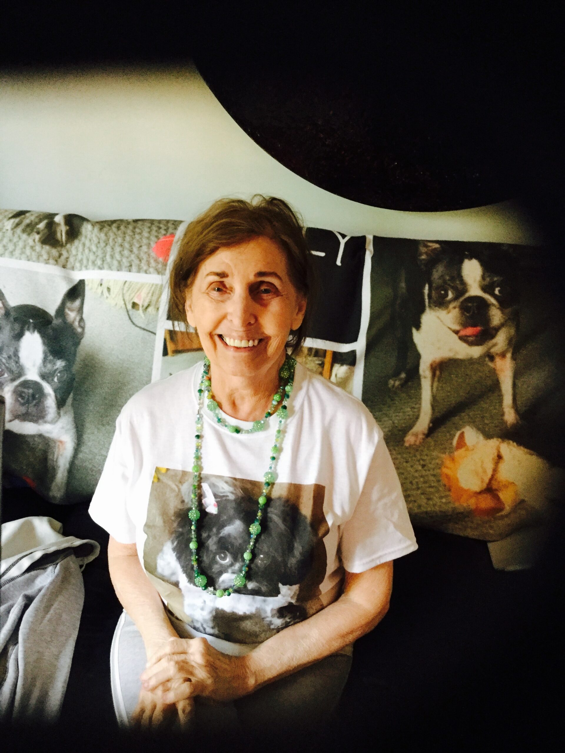 Smiling elderly woman in a white dog-printed shirt and green bead necklace, seated on a couch with large dog posters in the background.