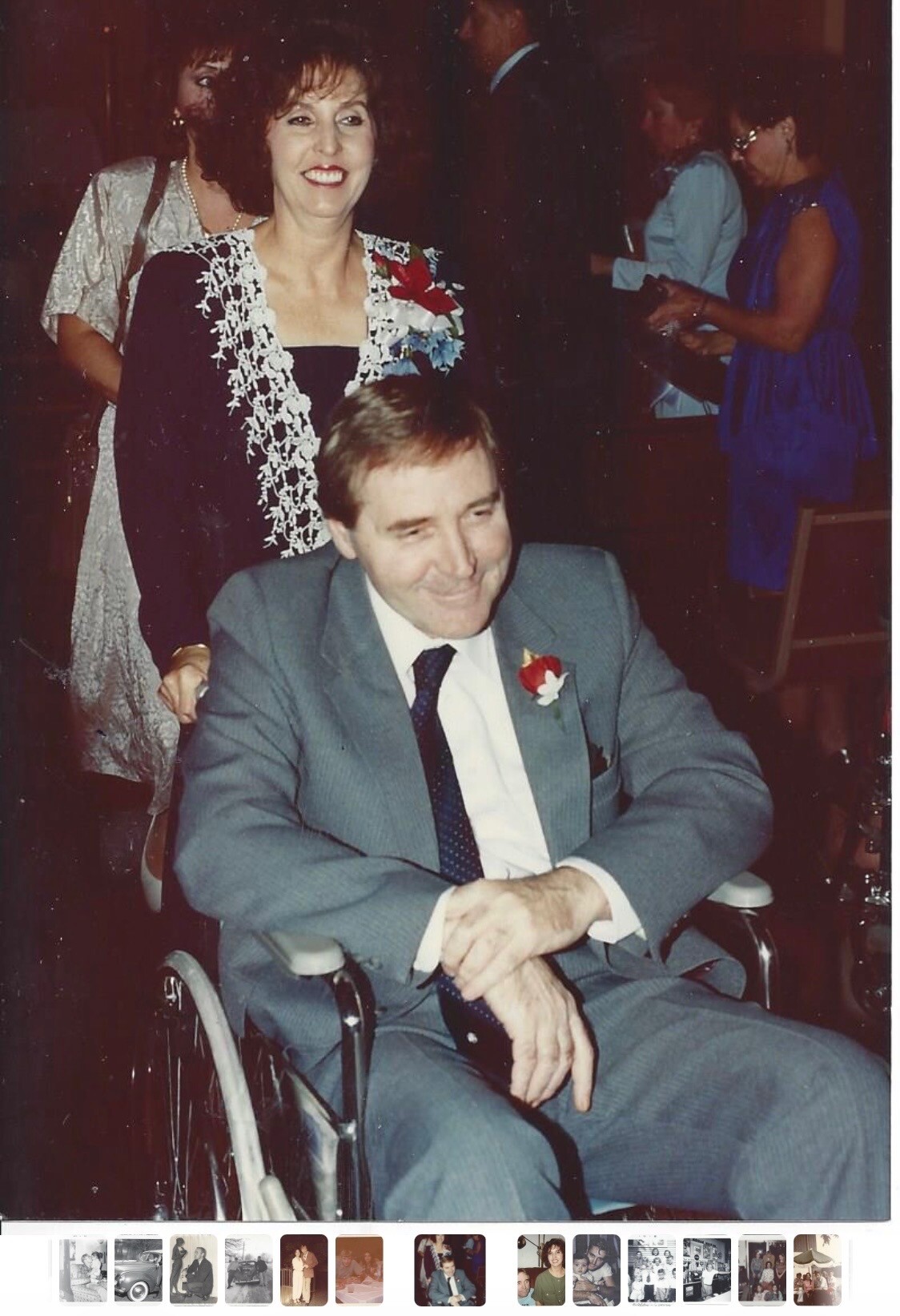 Smiling man in a gray suit in a wheelchair at a formal event, with a red boutonniere on his lapel; a woman stands behind him wearing a lacy outfit.