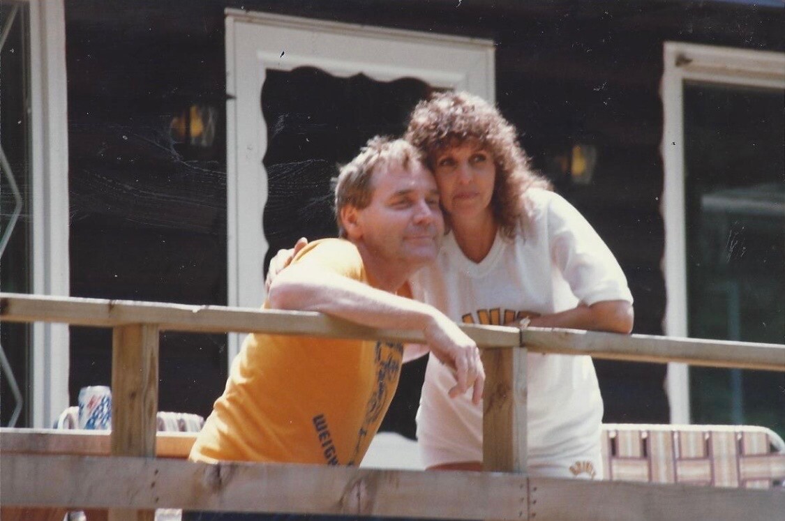 Man in an orange shirt rests his arms on a wooden railing while a curly-haired woman in a white shirt stands beside him on a deck outside a house.