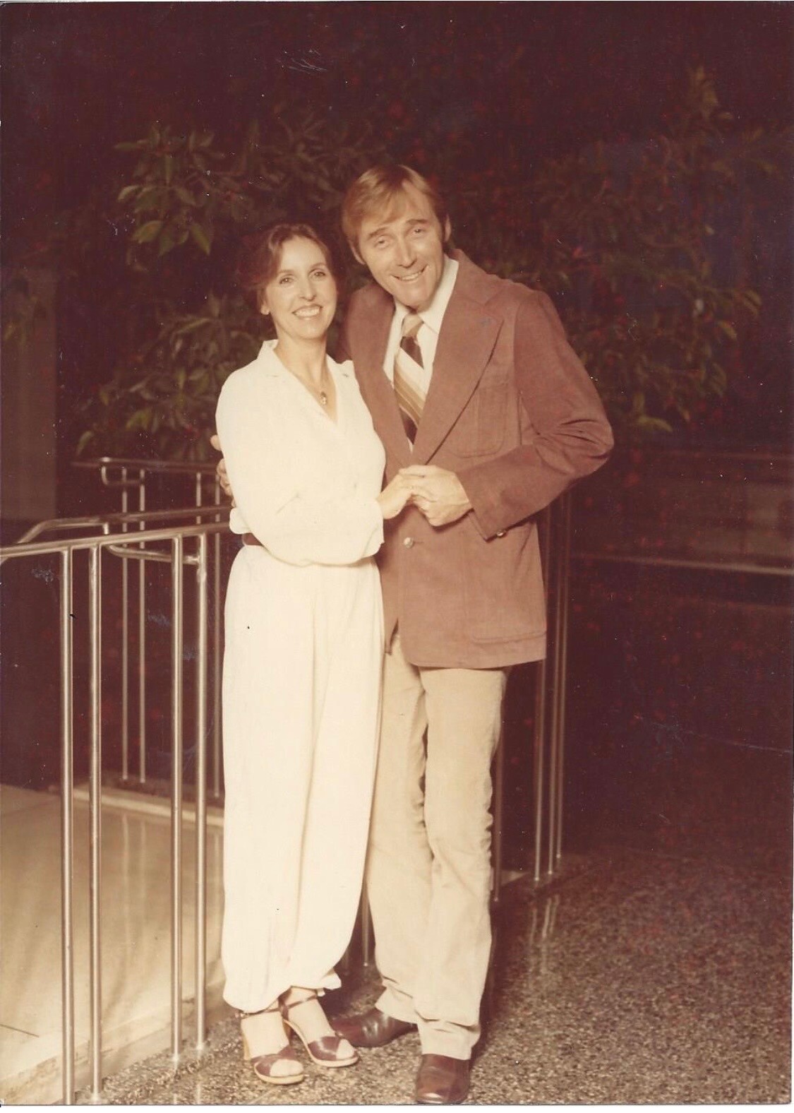 Smiling couple standing close together, arms linked; woman in white outfit and sandals, man in a brown blazer and striped tie.