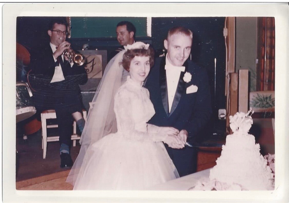 Bride and groom in formal wedding attire cutting their cake at the reception, with a band playing in the background.
