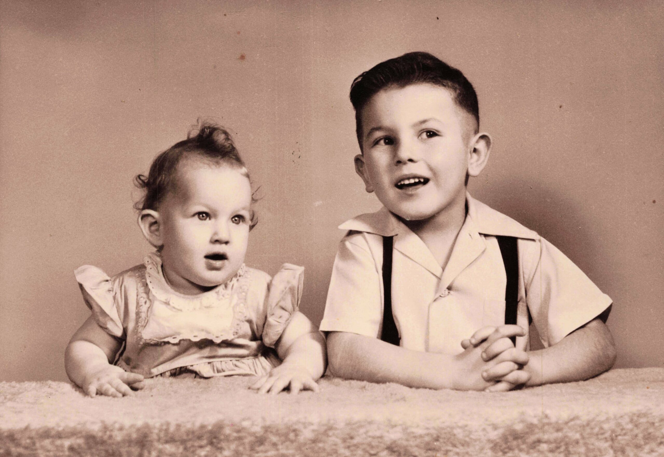 Two young siblings sit side by side on a rug against a plain backdrop; a baby girl in a lacy dress on the left and a boy in a collared shirt with suspenders on the right, looking off-camera.