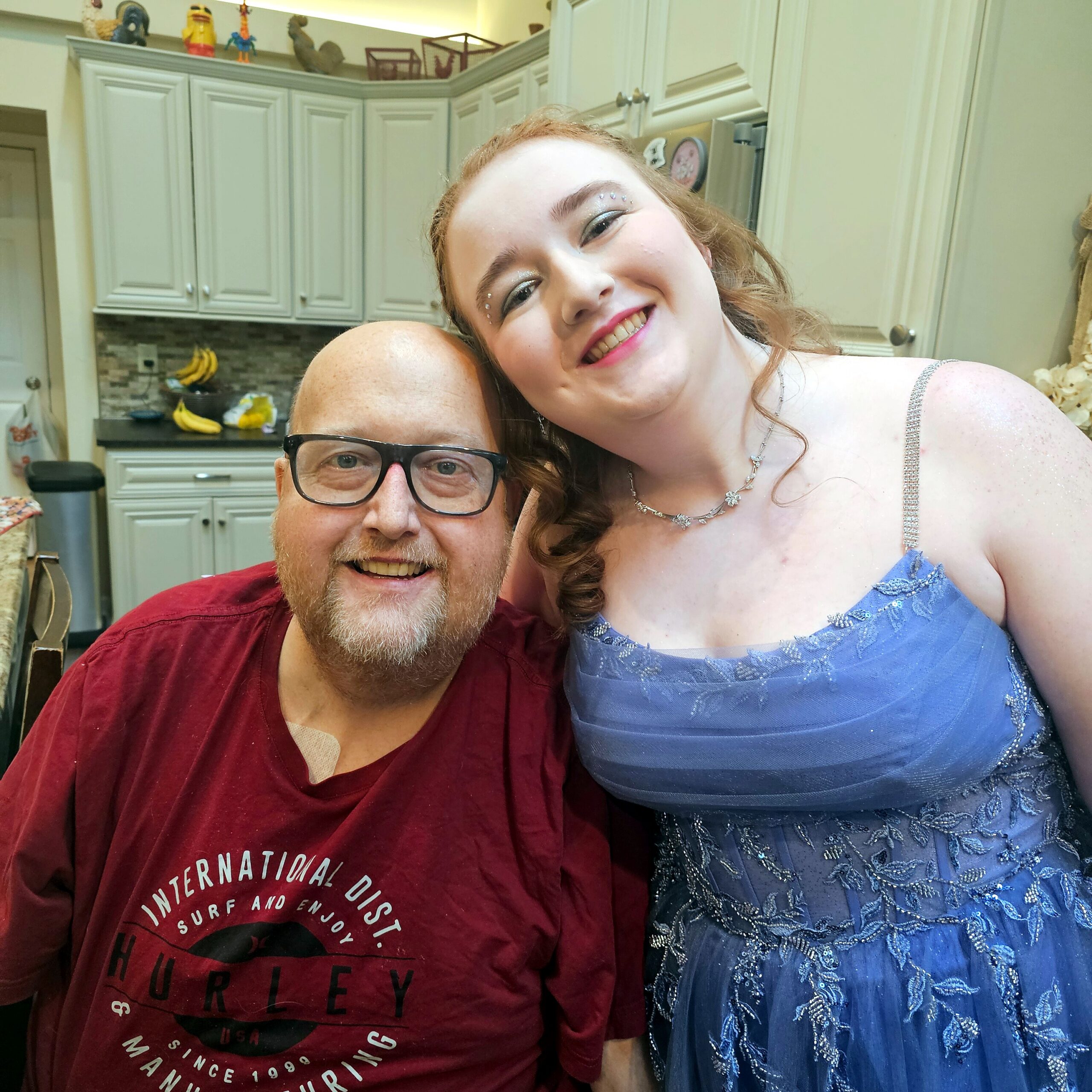 Smiling couple posing for a selfie in a bright kitchen, woman in a blue dress leaning on a man in a red shirt with glasses.