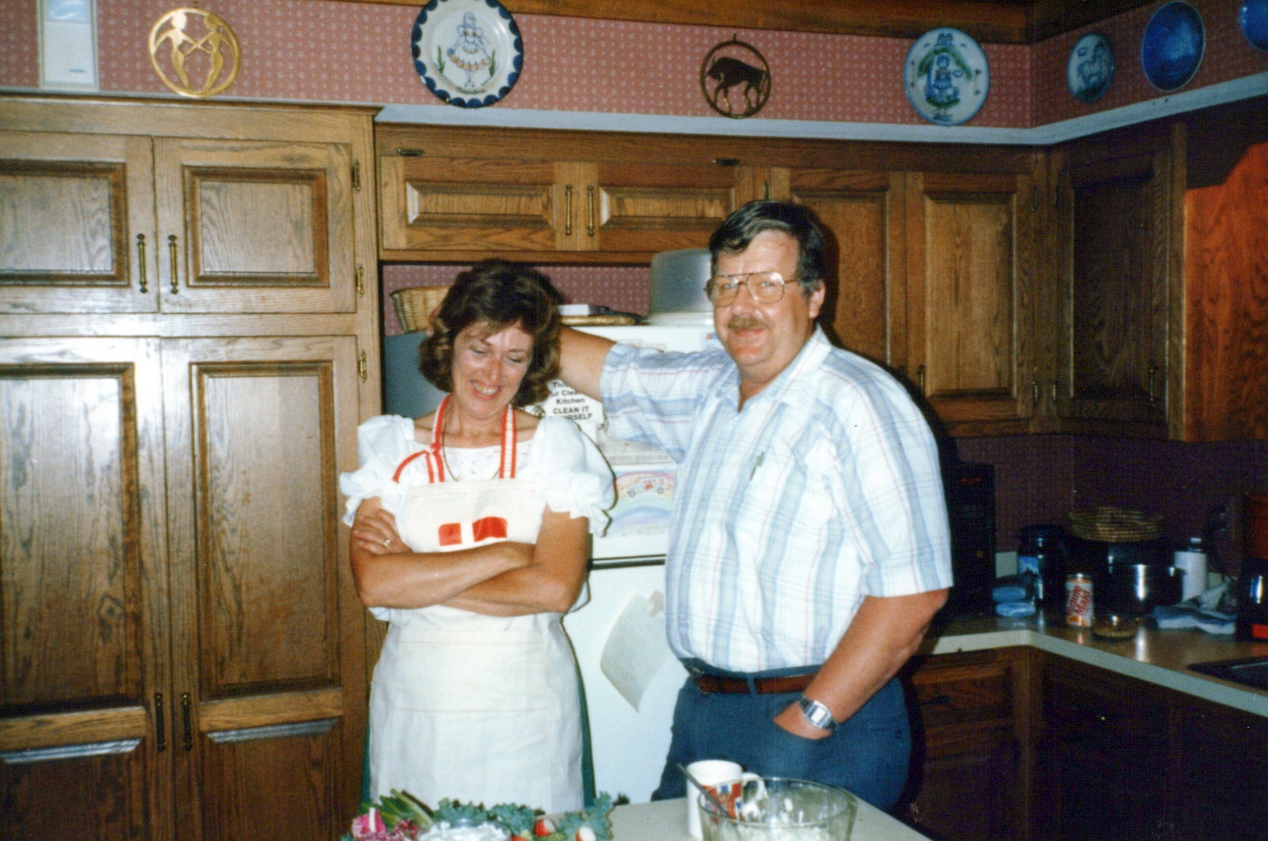 Smiling couple stands in a wood-panel kitchen; woman wears a white apron, man in a plaid shirt with glasses, posing together.