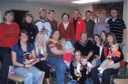 Large group of family and friends posing for a group photo in a living room, with children and babies among them.