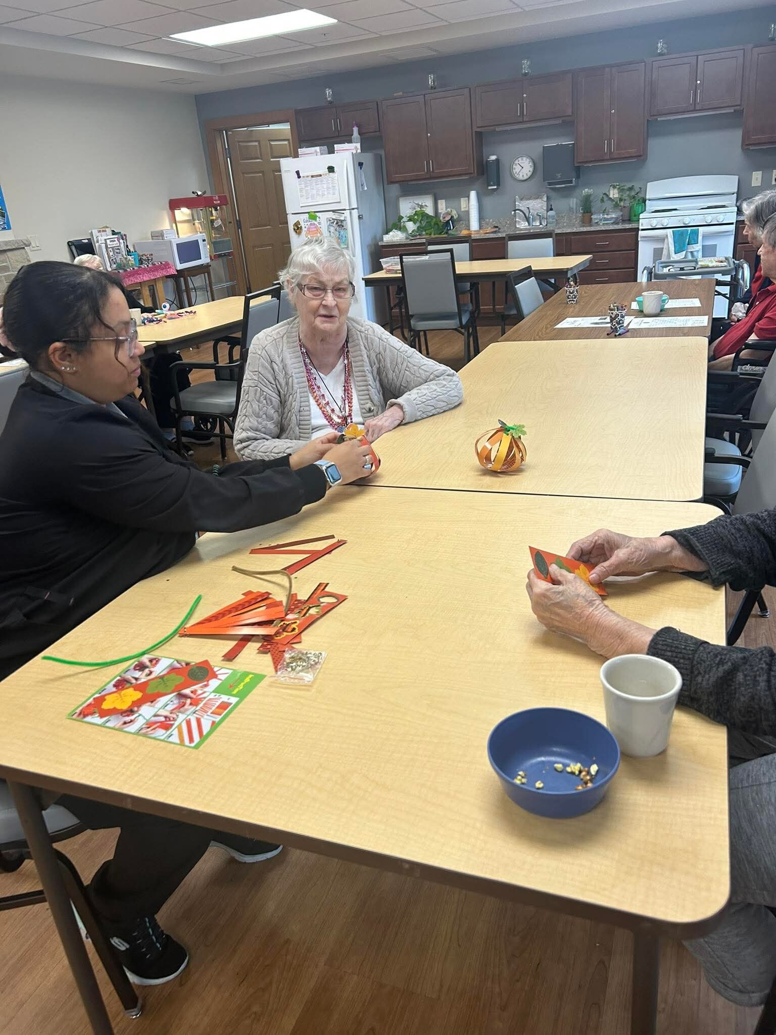 Group of seniors seated around a long table, making autumn paper crafts with colorful strips in a community room kitchen area in the background.