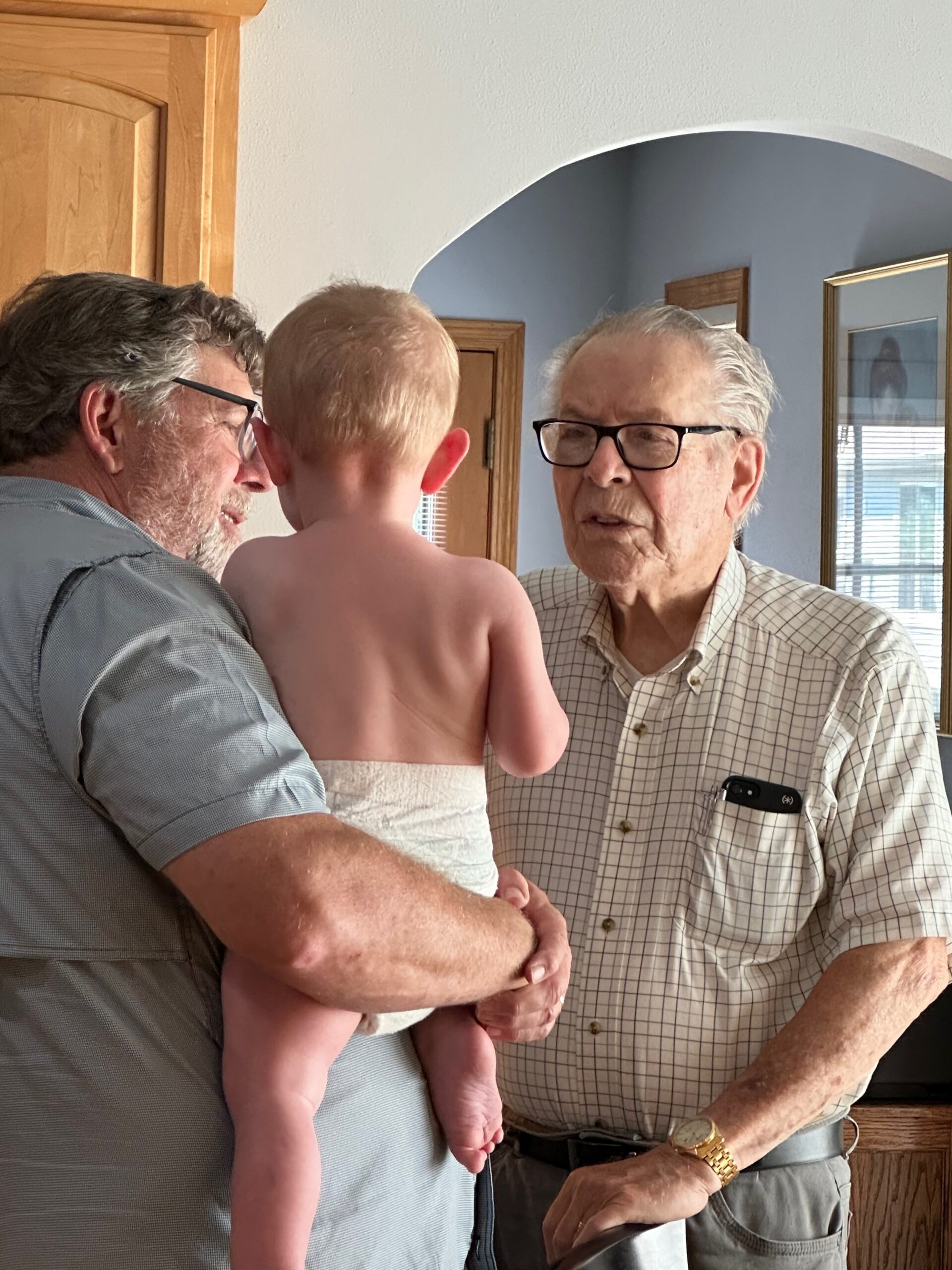 Two elderly men hold a naked baby between them inside a home, standing near a wooden cabinet and archway.
