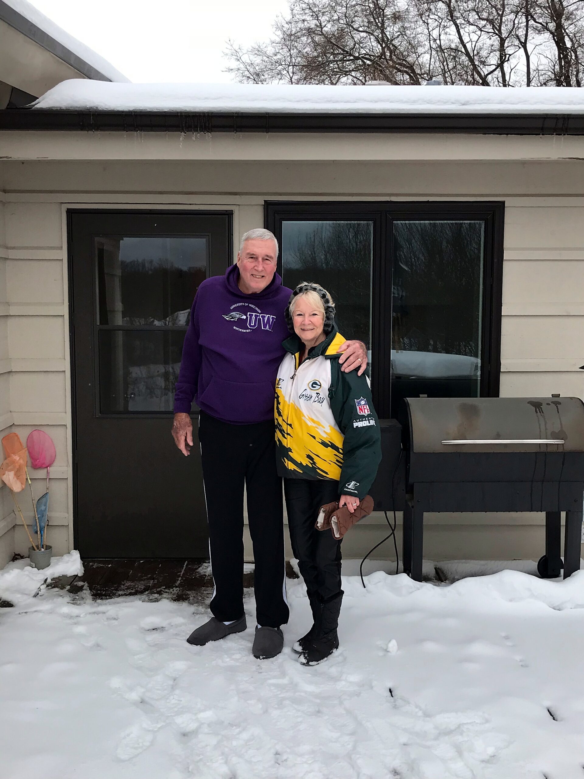 Two elderly adults pose together on a snowy patio in front of a house, arms around each other, wearing warm jackets and smiles.