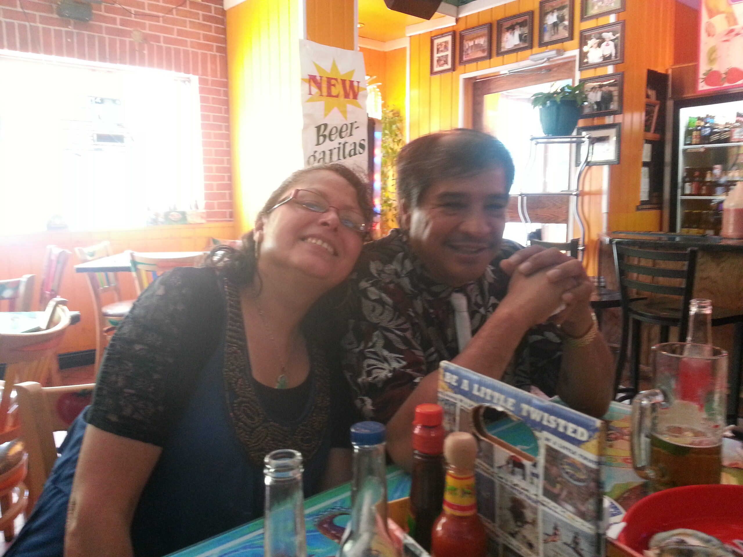 Two friends smiling at a crowded casual restaurant table, with a wall of photos and a bright yellow sign in the background.