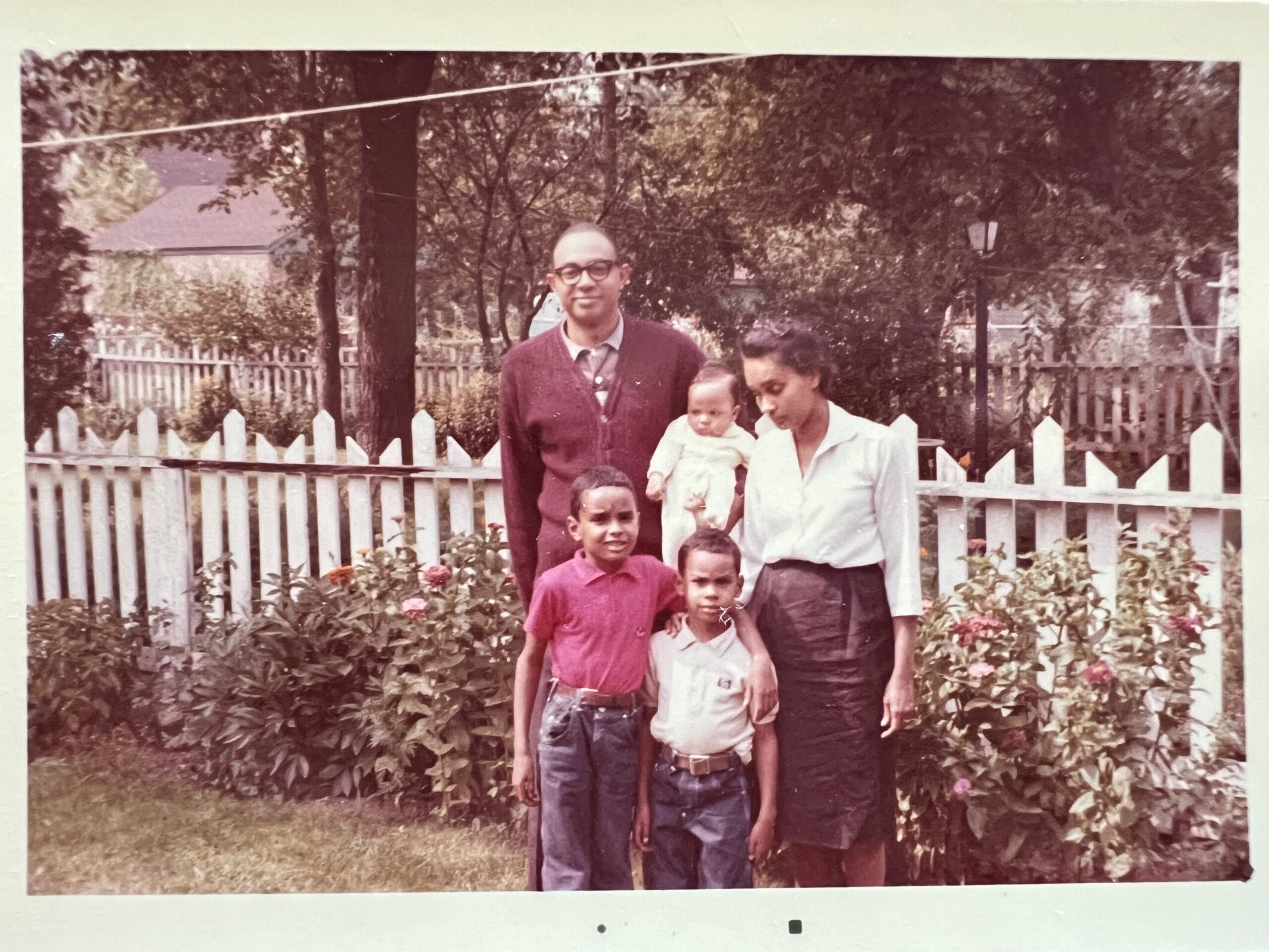 A family of five outdoors in a garden, posing in front of a white picket fence with plants and flowers nearby.