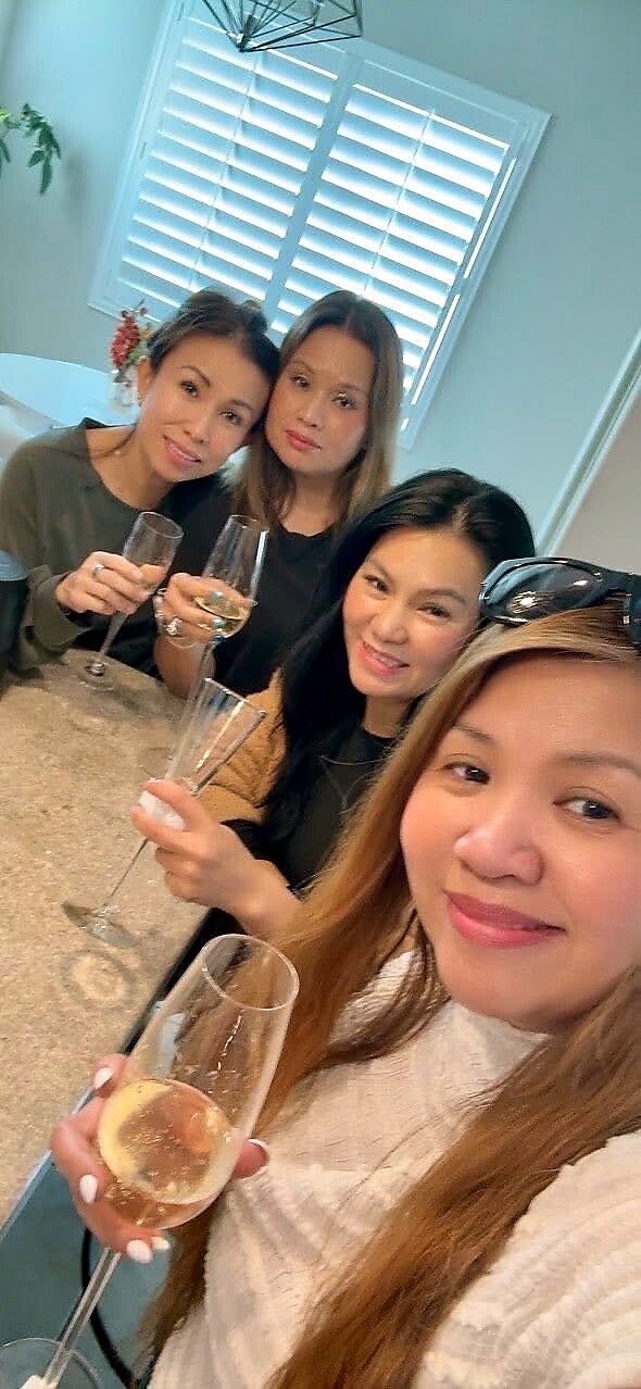 Four women pose for a selfie, toasting with champagne flutes in a bright kitchen dining area.