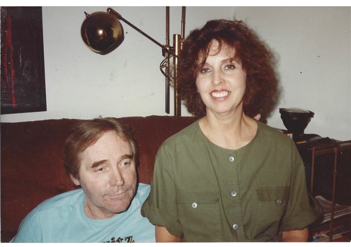 Smiling woman in an olive-green shirt stands beside a seated man in a light blue shirt on a brown couch; a floor lamp is visible behind them inside a living room.